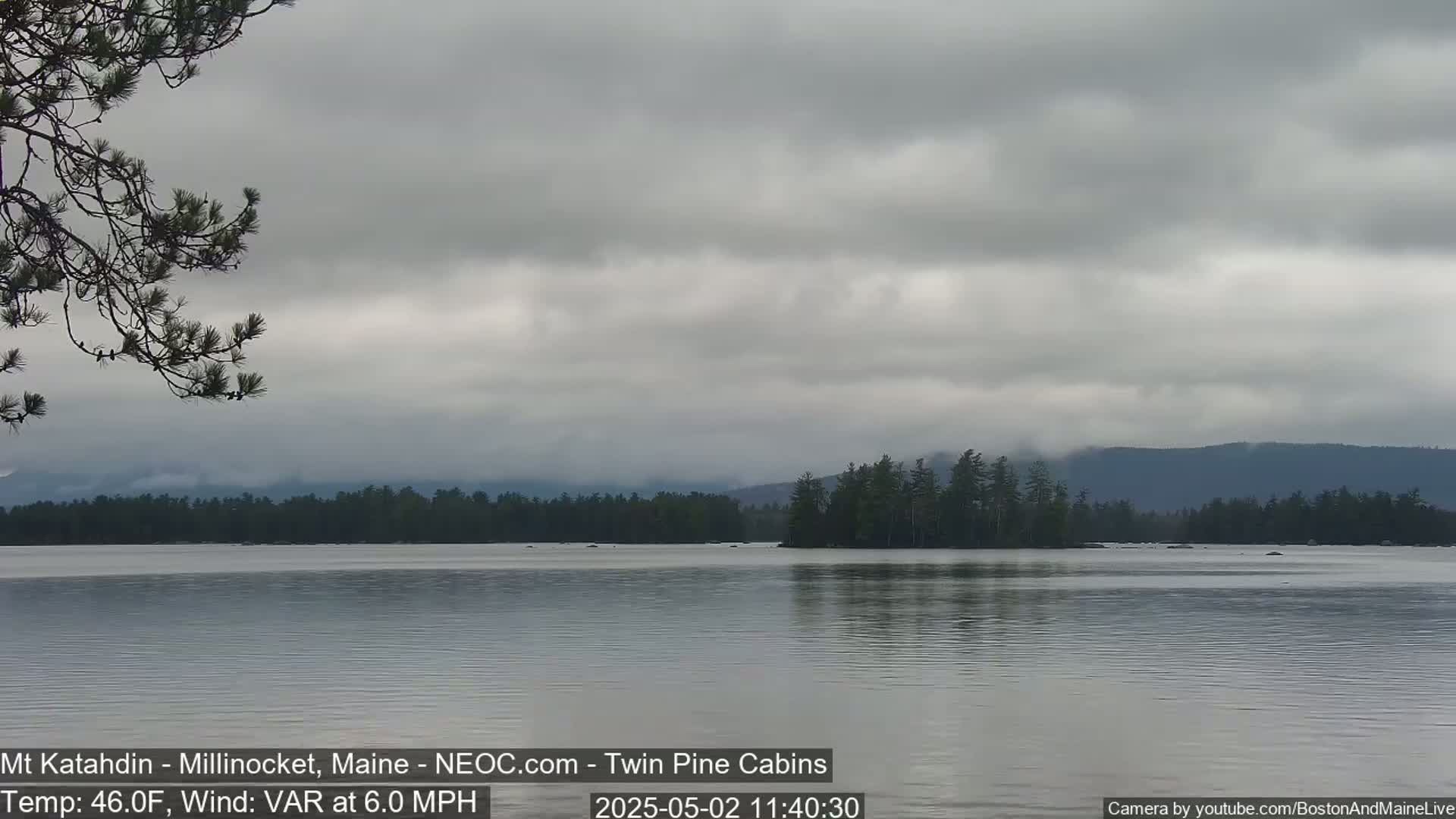 A calm lake under an overcast sky is surrounded by dark green trees and distant mountains.