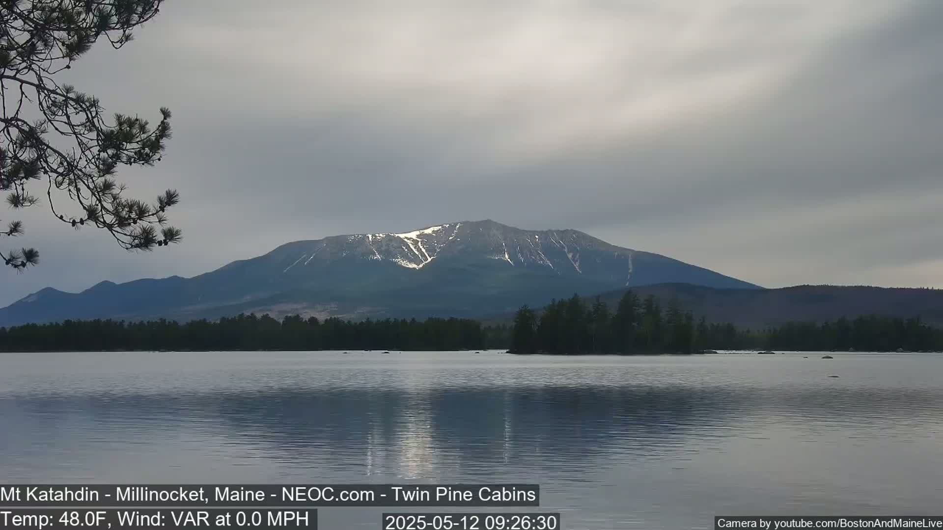 A calm lake reflects a snow-capped mountain under an overcast sky.