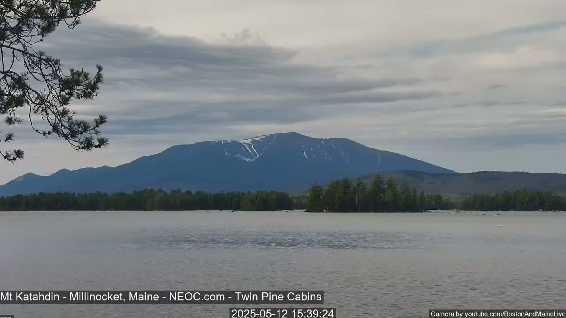 A calm lake reflects a mountain range under a partly cloudy sky, with some snow still visible on the mountain's peaks.