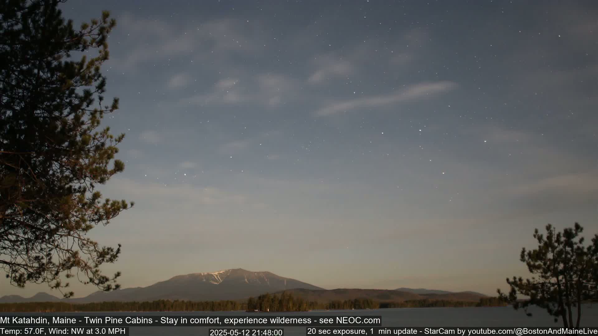 A starry night sky with some clouds above a snow-capped mountain range and dark silhouettes of pine trees, under a calm, cool breeze.