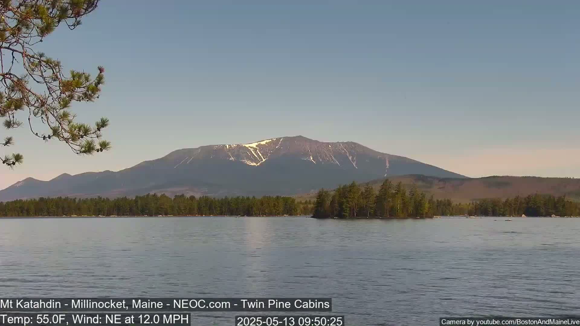 A calm lake with a forested shoreline reflects a mountain range with some snow on its peaks under a clear sky, with a northeast wind at 12 mph and a temperature of 55°F.