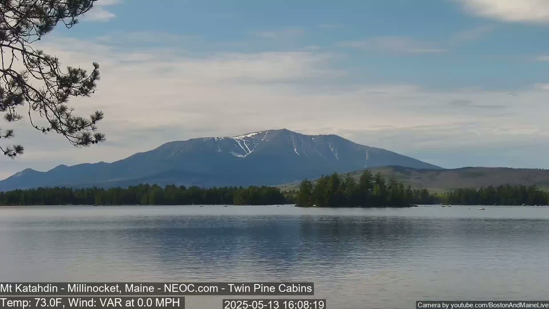 A calm lake reflects a mountain range with patches of snow under a mostly clear sky.