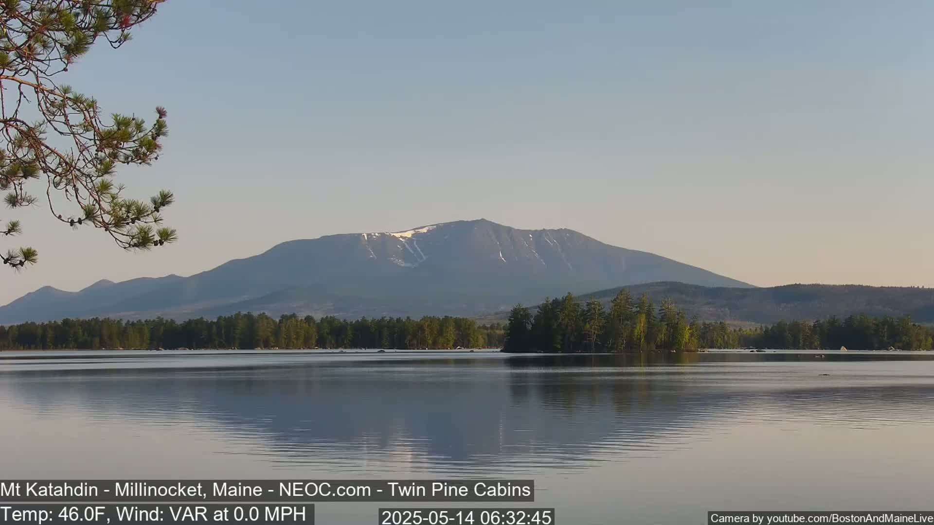 A calm lake reflects a mountain range with patches of snow under a clear sky.