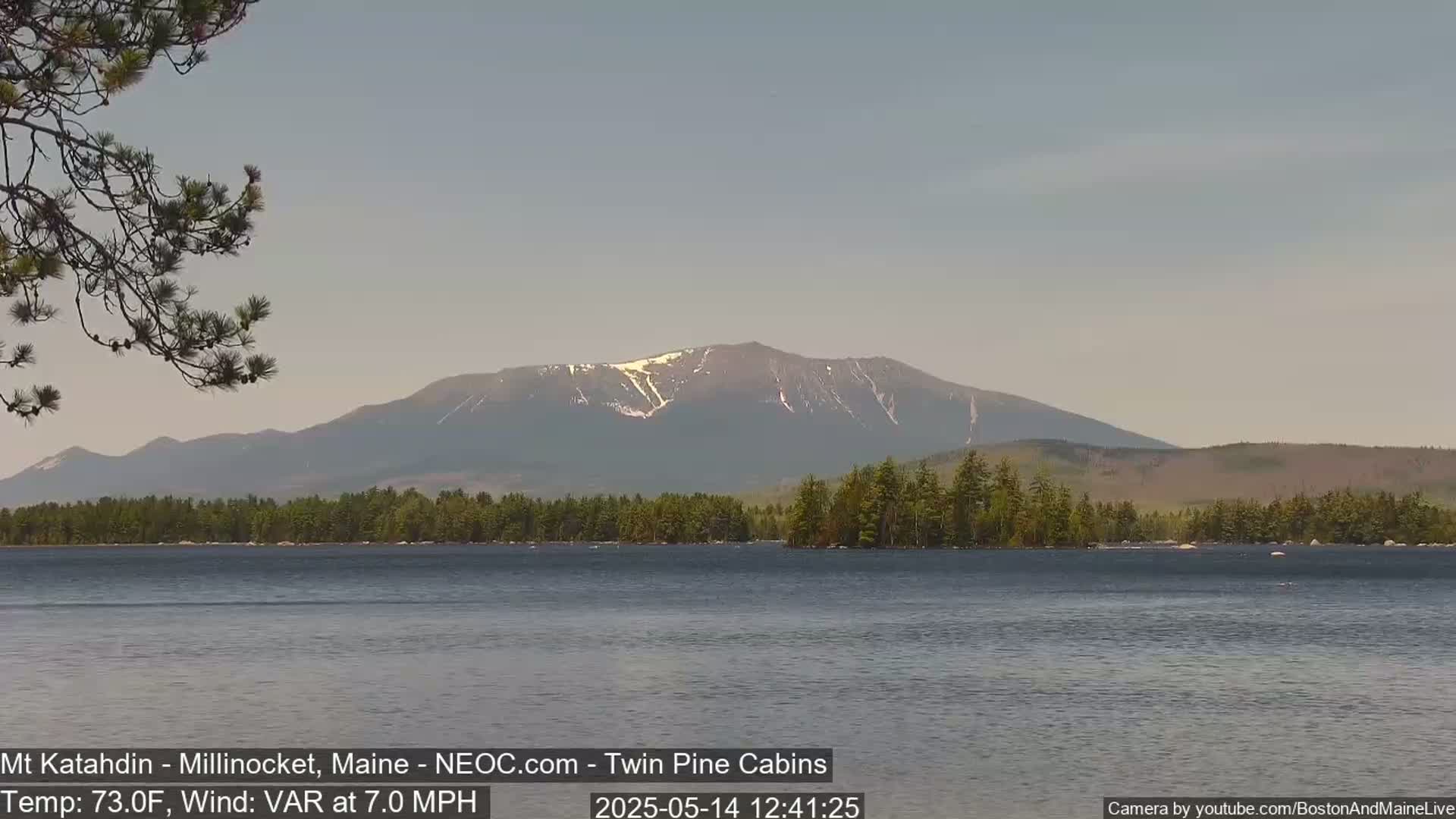 A calm lake reflects a mountain range with some remaining snow, under a clear, light-grey sky.