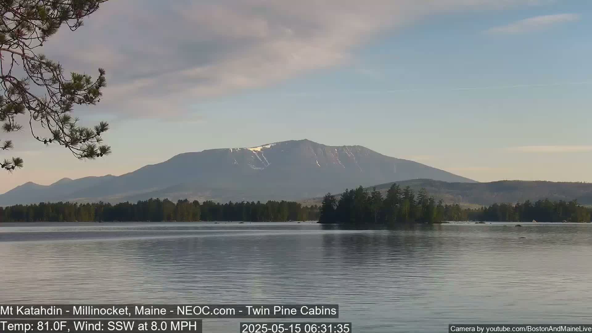 A calm lake reflects a mountain range under a partly cloudy sky, with a slight breeze from the south-southwest.