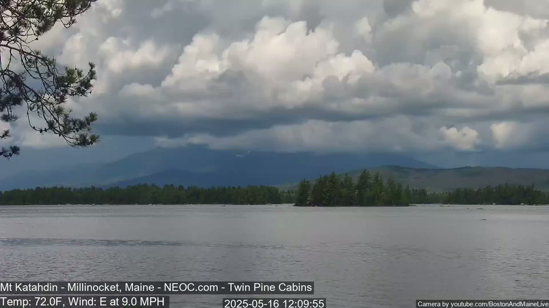 A calm lake reflects a cloudy sky and distant mountains under partly cloudy conditions with an easterly wind.