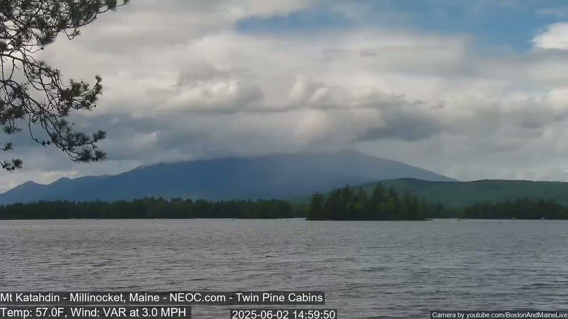 A calm lake reflects a mountain range under a cloudy sky with a 3.0 mph variable wind and a temperature of 57°F.