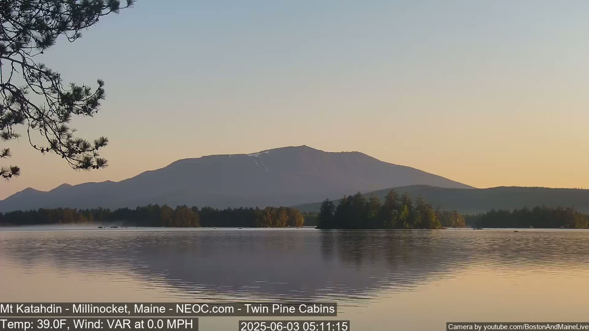 A calm lake reflects a mountain range under a pale sunrise sky.