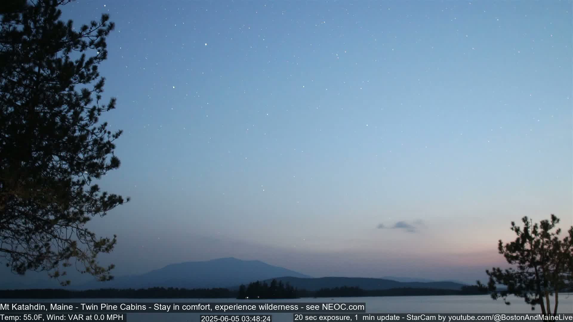 Under a twilight sky dotted with stars, a calm body of water reflects the silhouette of distant mountains and trees.