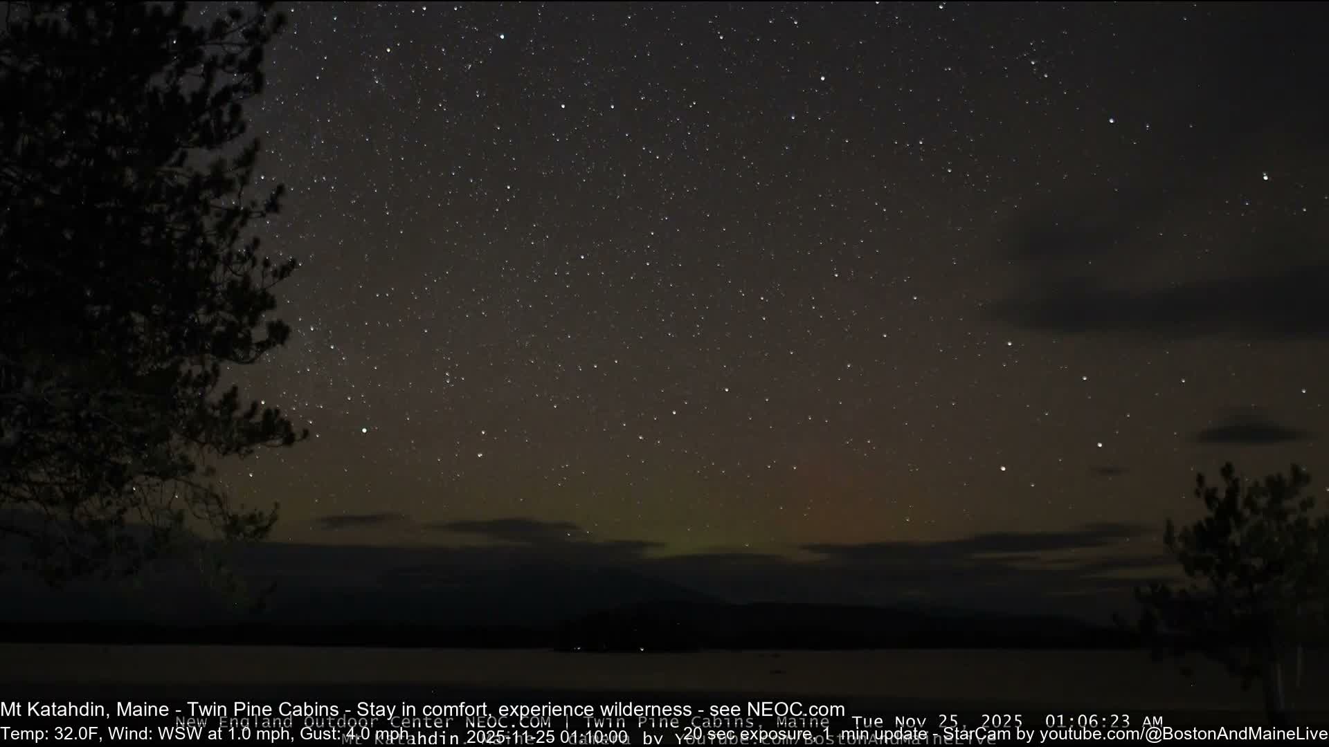 A clear, star-filled night sky with a faint green and orange auroral glow on the horizon stretches above a dark body of water and silhouetted trees, with conditions appearing calm and very cold.