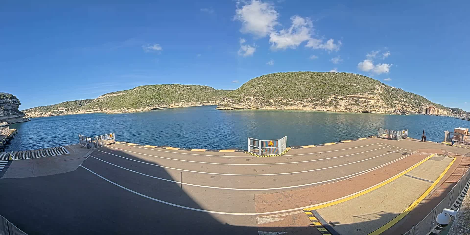 A ferry terminal with a paved roadway and metal gates overlooks a calm body of water bordered by green, rocky hills under a mostly sunny sky.