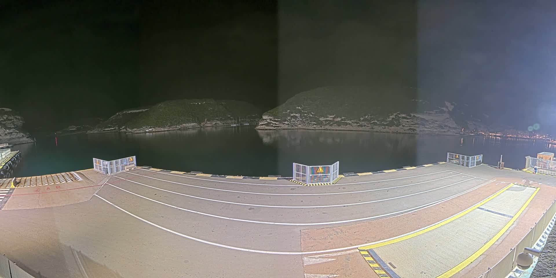 A nighttime view of a ferry terminal's empty loading dock, with calm water reflecting the dark, hilly shoreline.