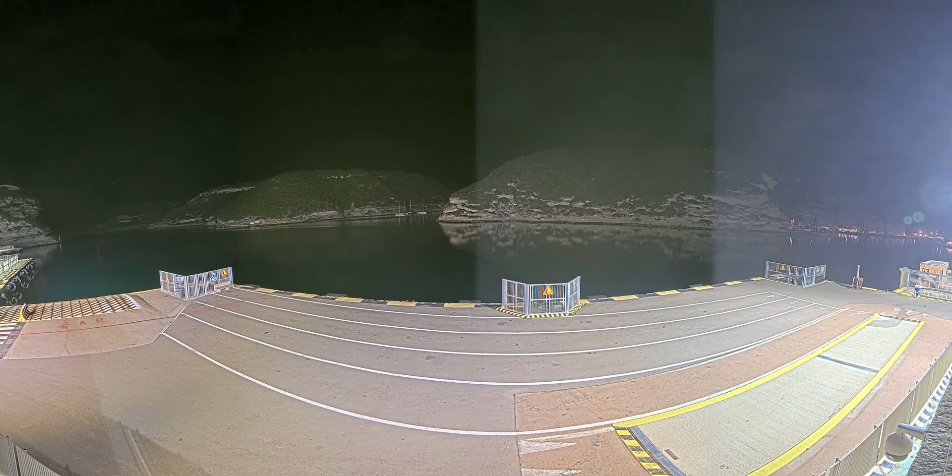 An empty ferry dock at night, illuminated by artificial lights, overlooks a calm body of water nestled between dark hills.