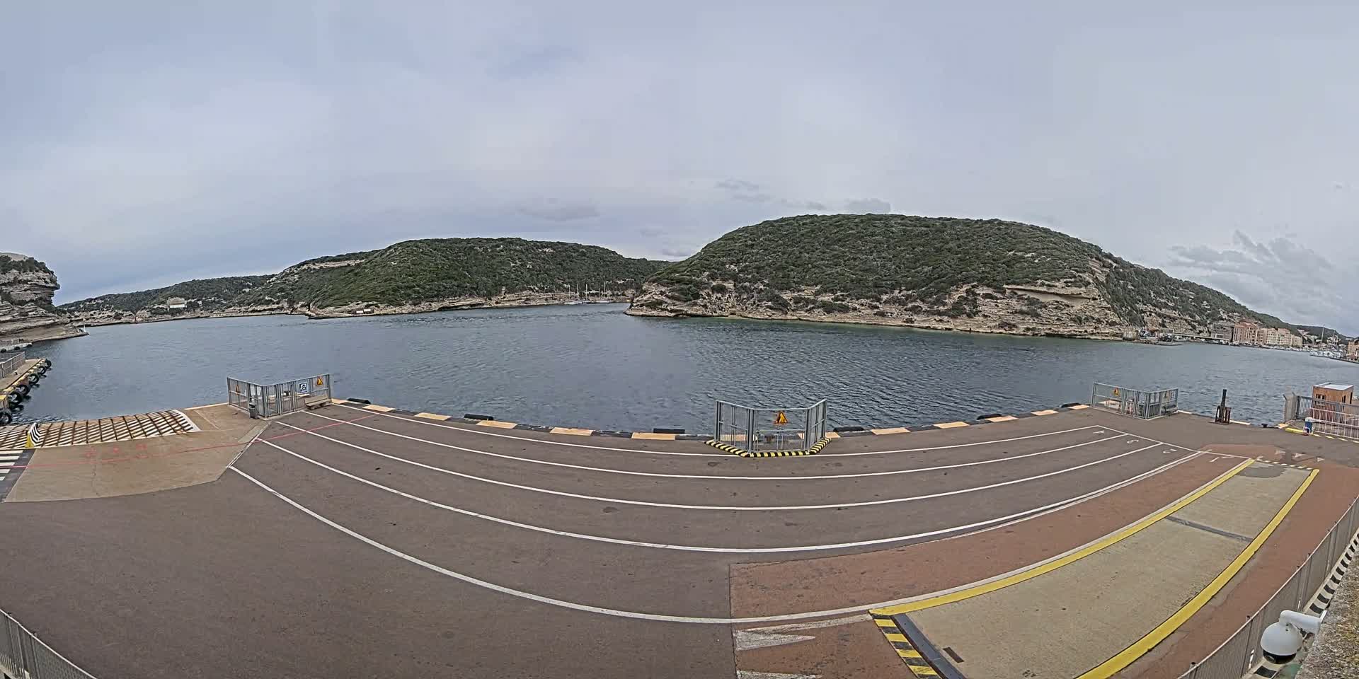 An empty ferry dock with marked lanes overlooks a calm body of water surrounded by green, hilly terrain under an overcast sky.