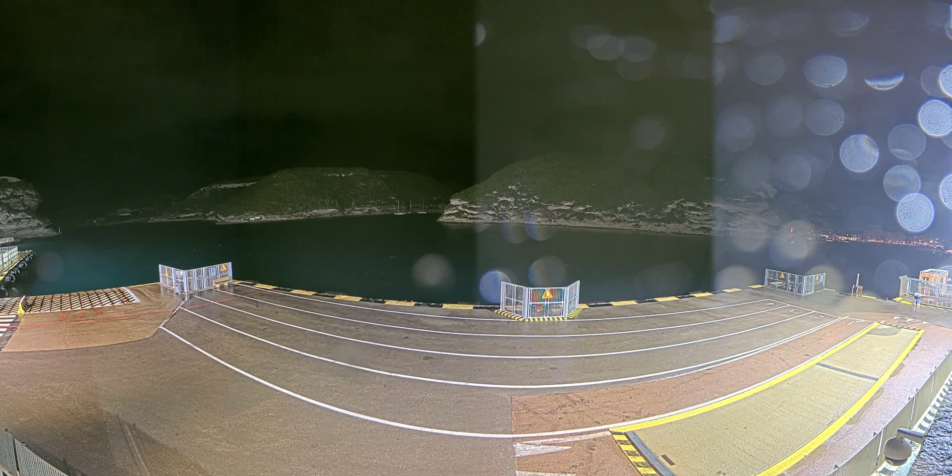 A nighttime view of a rain-slicked ferry dock, with calm water and dark hills visible in the background.