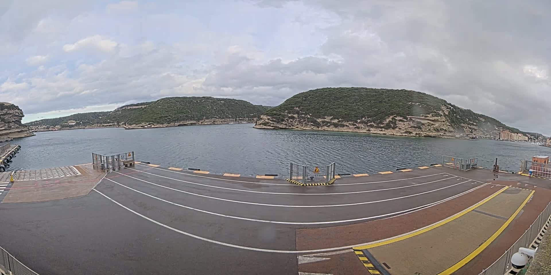 A wet ferry terminal with marked lanes overlooks a calm bay surrounded by green, hilly land under a cloudy sky.