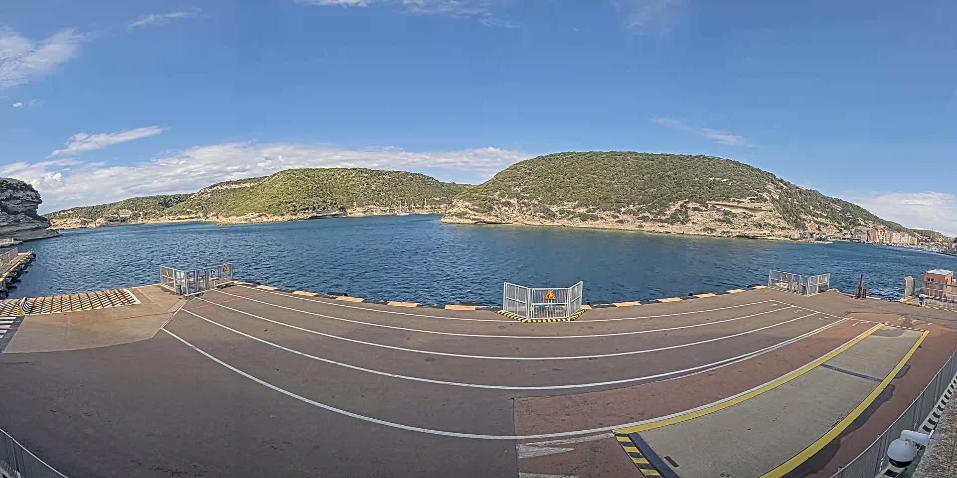 A ferry dock with a calm body of water and green hills under a mostly sunny sky.