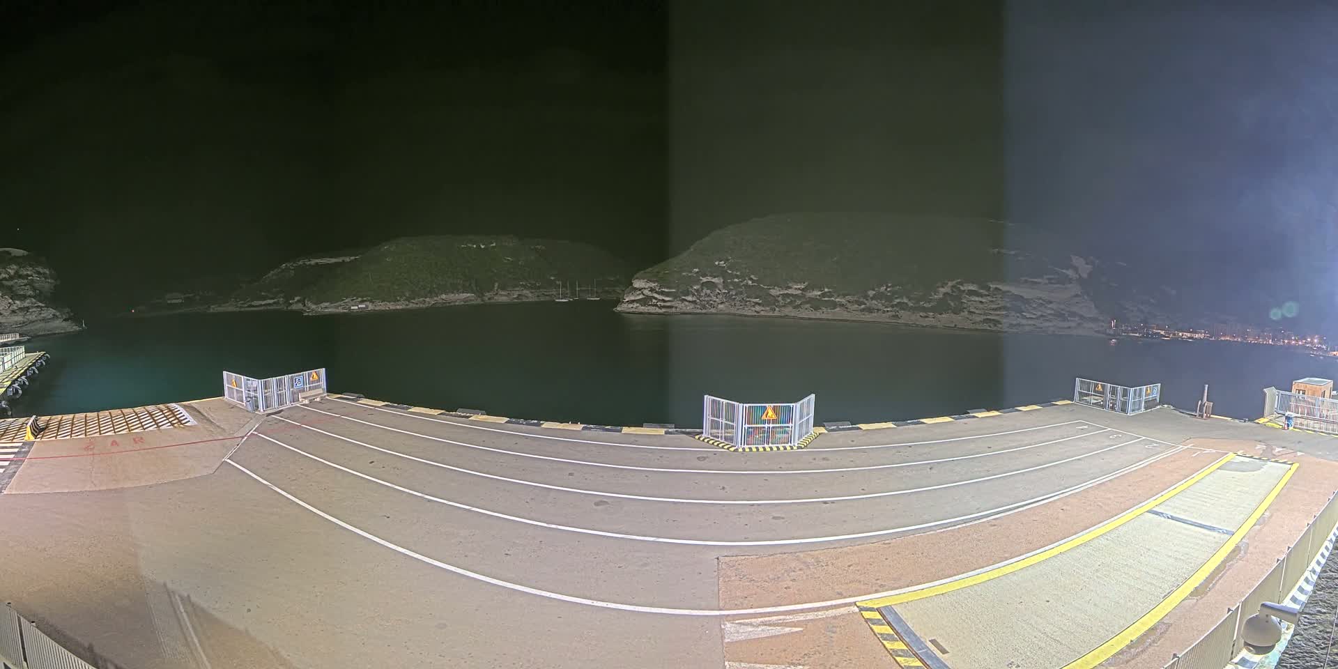 An empty ferry dock at night is illuminated, overlooking a dark body of water surrounded by hills under a clear, dark sky.