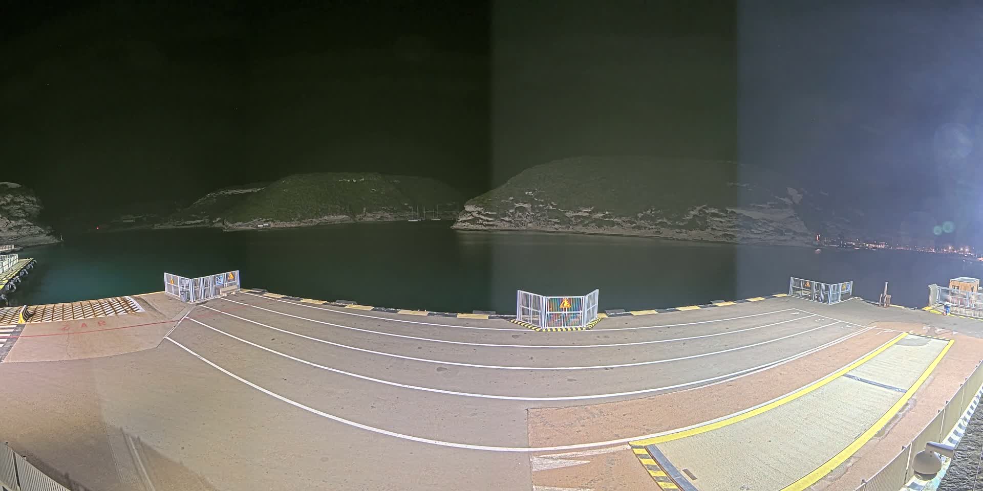 An empty ferry dock at night, illuminated, overlooks a dark calm body of water surrounded by land.