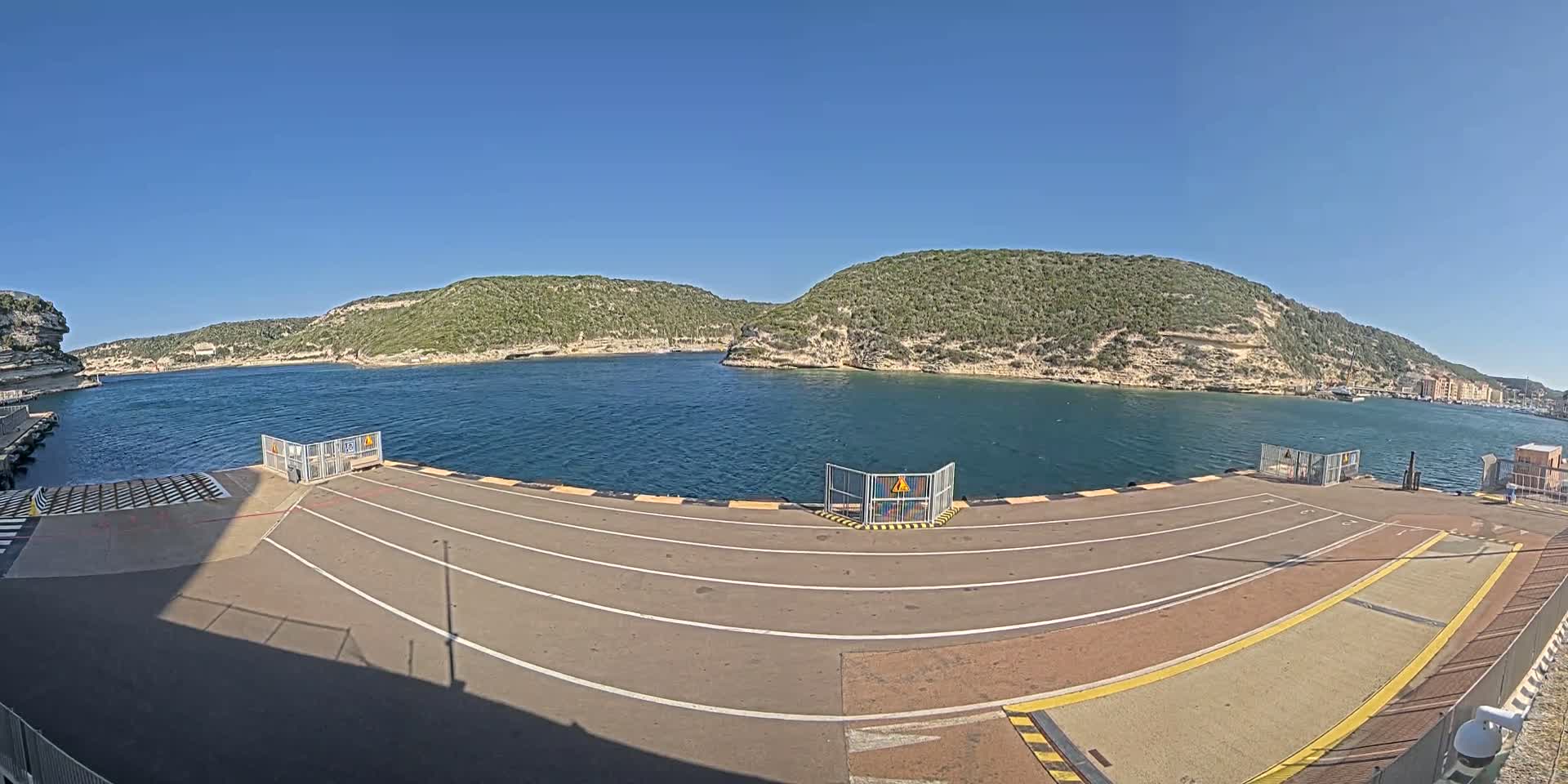 A ferry dock with marked lanes overlooks calm blue water and green, rocky hills under a clear blue sky.