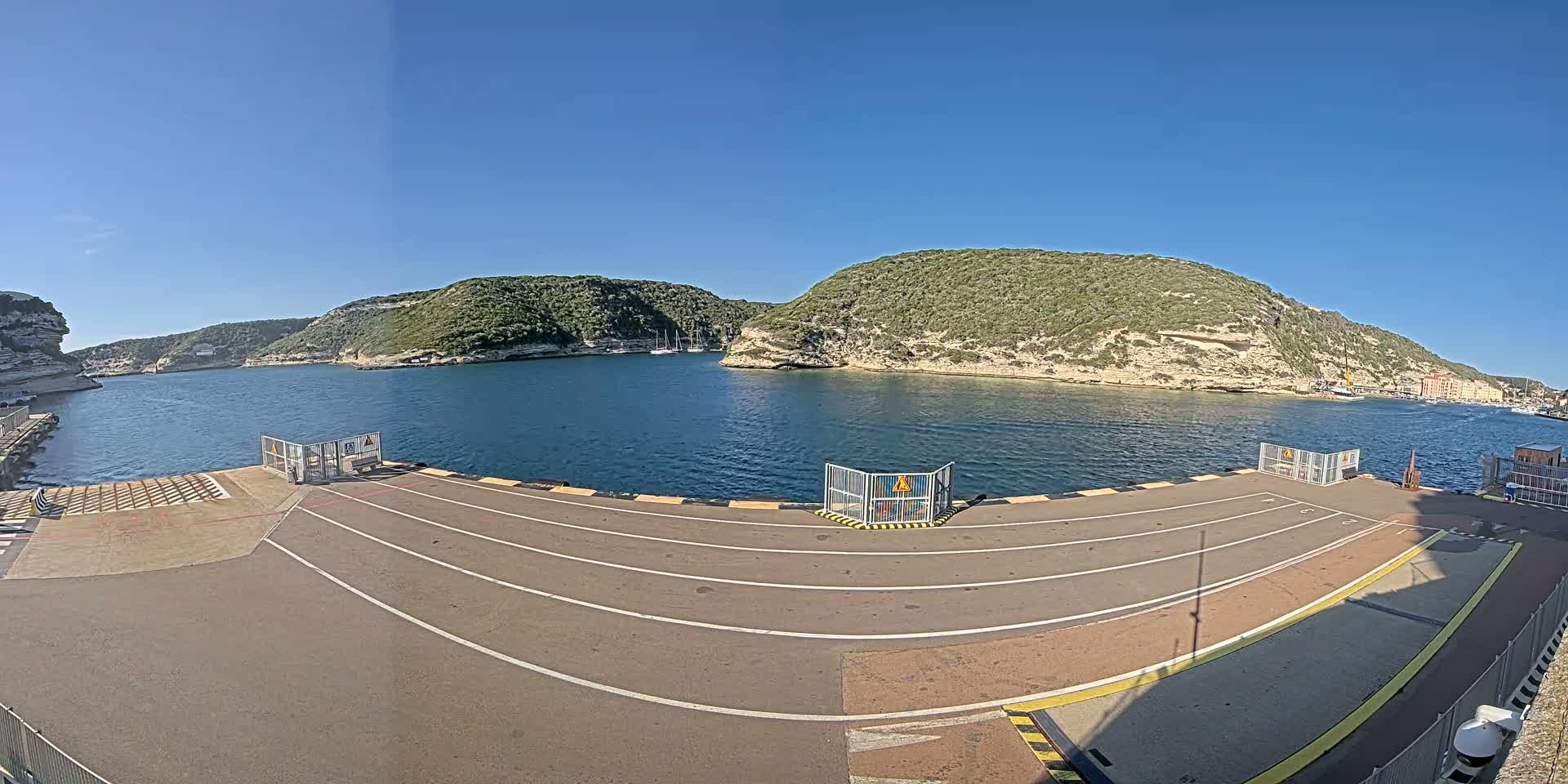 An empty ferry terminal with marked lanes overlooks a calm bay surrounded by green, rocky hills under a clear blue sky.
