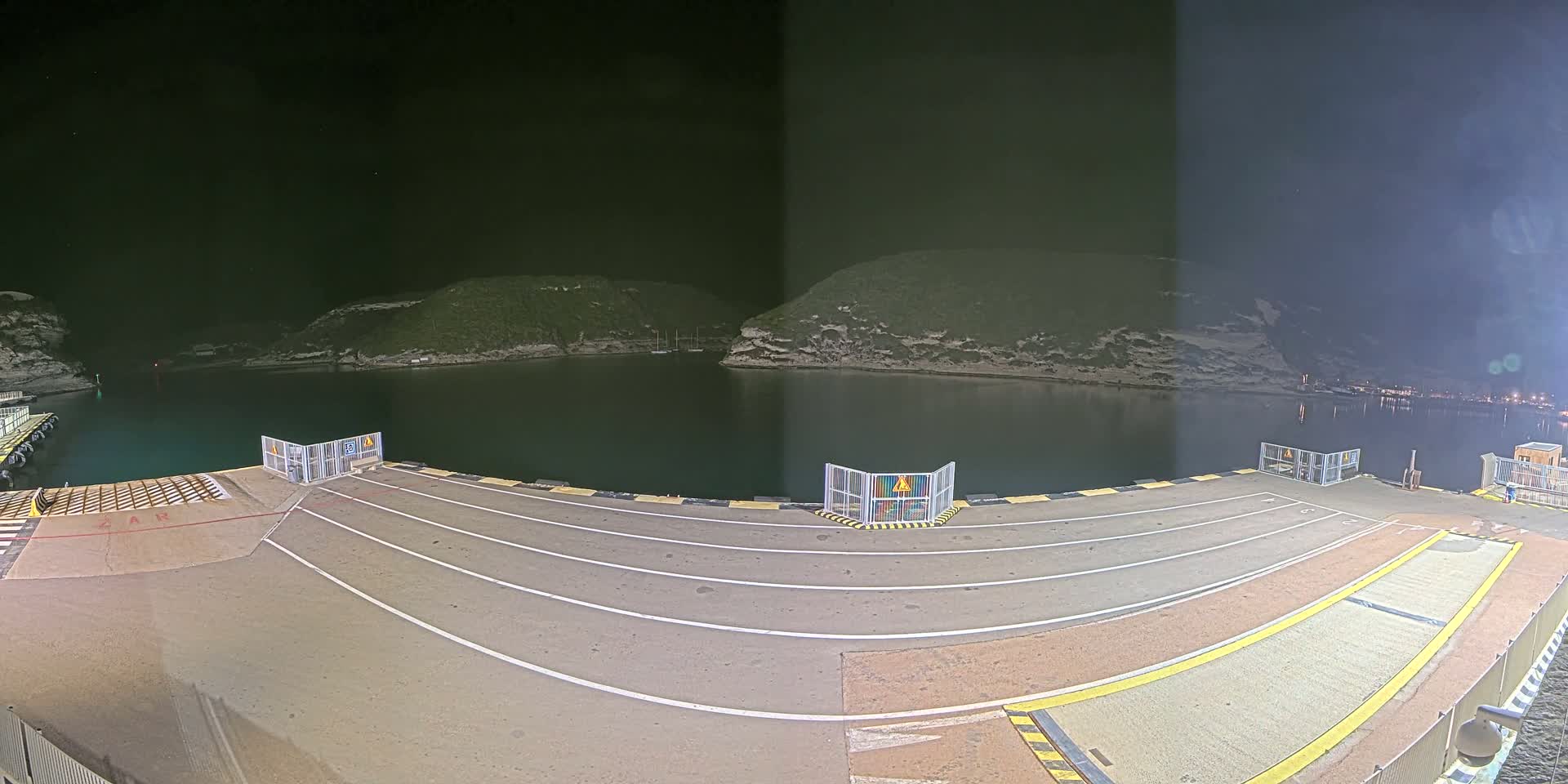 A nighttime view of a ferry dock with calm water and hills in the background under a clear, dark sky.