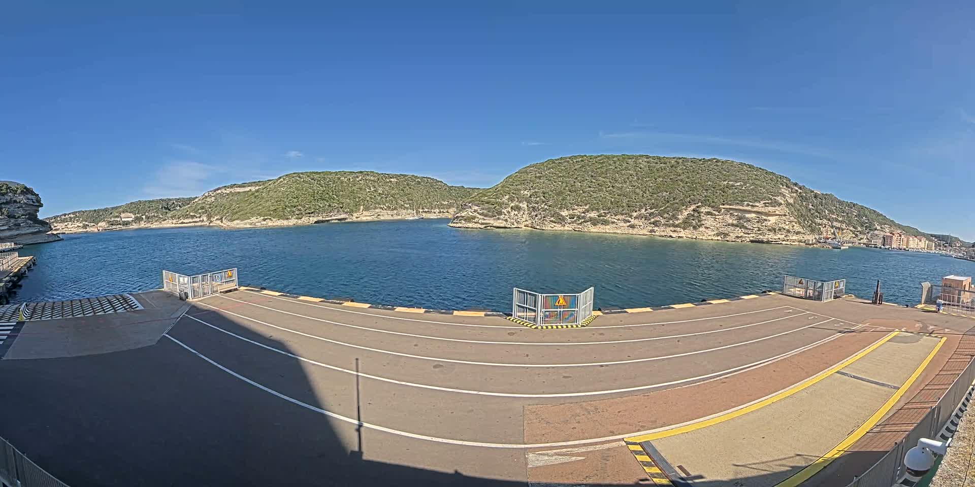 A ferry terminal's empty roadway overlooks a calm bay surrounded by green, rocky hills under a clear blue sky.