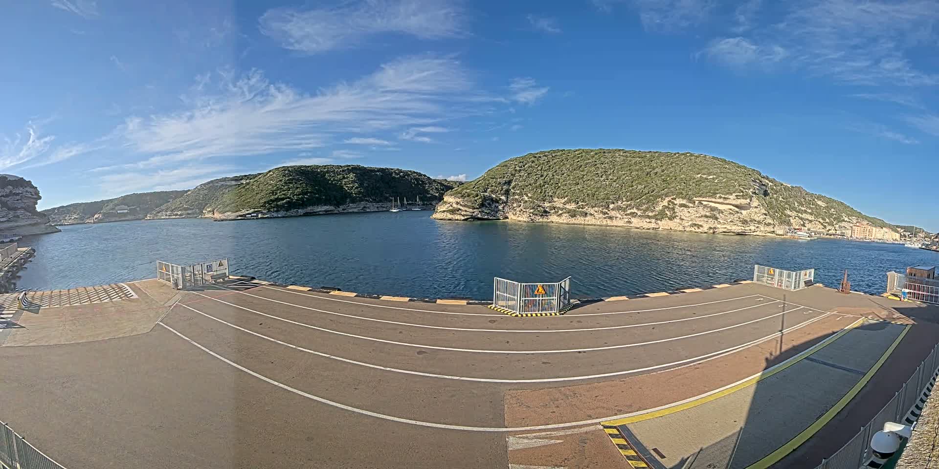 A ferry terminal with marked lanes overlooks a calm bay surrounded by rocky, green hills under a mostly sunny sky.
