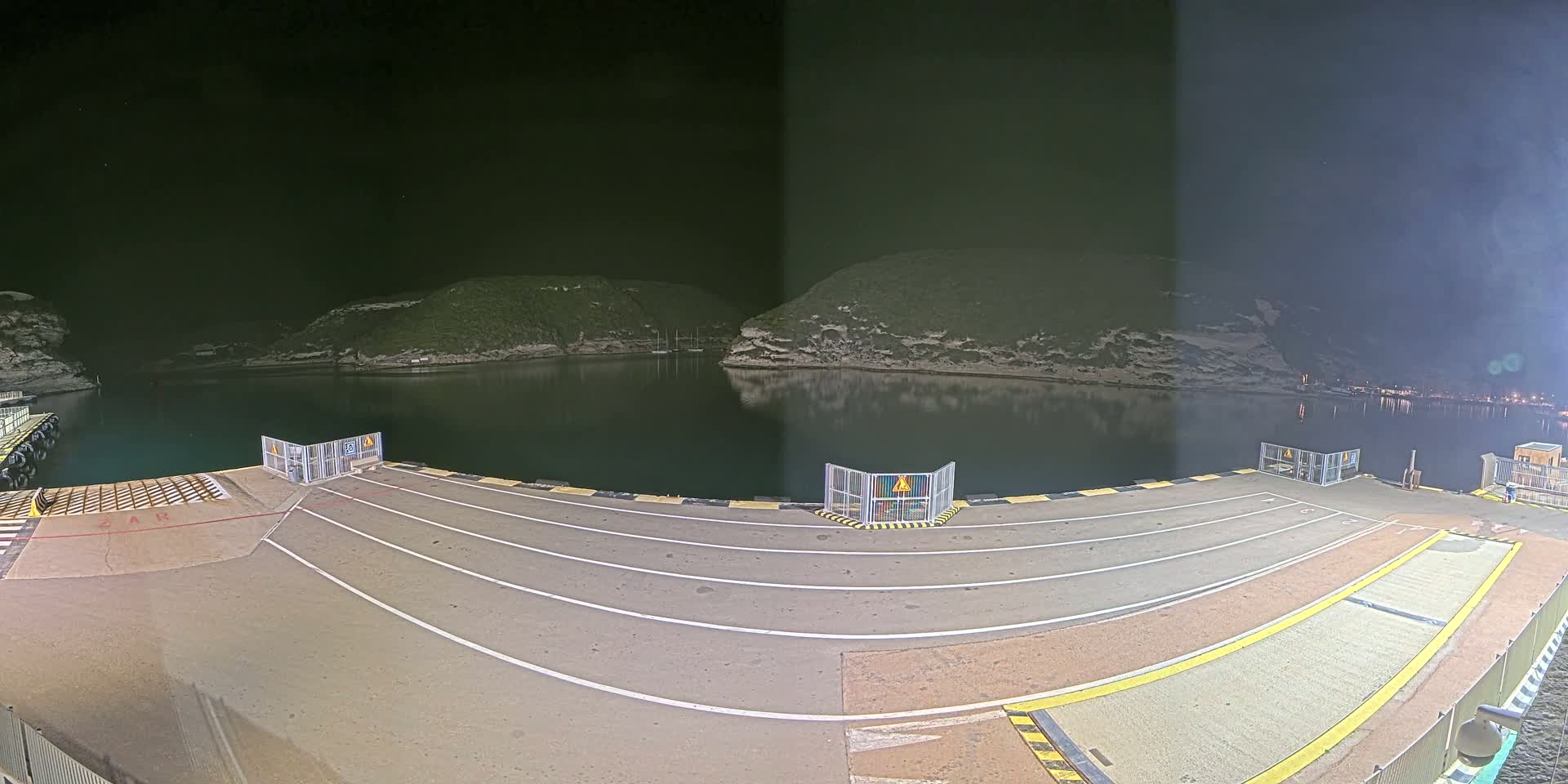 An empty ferry dock at night, illuminated, overlooks a calm bay nestled between dark hills under a clear, dark sky.