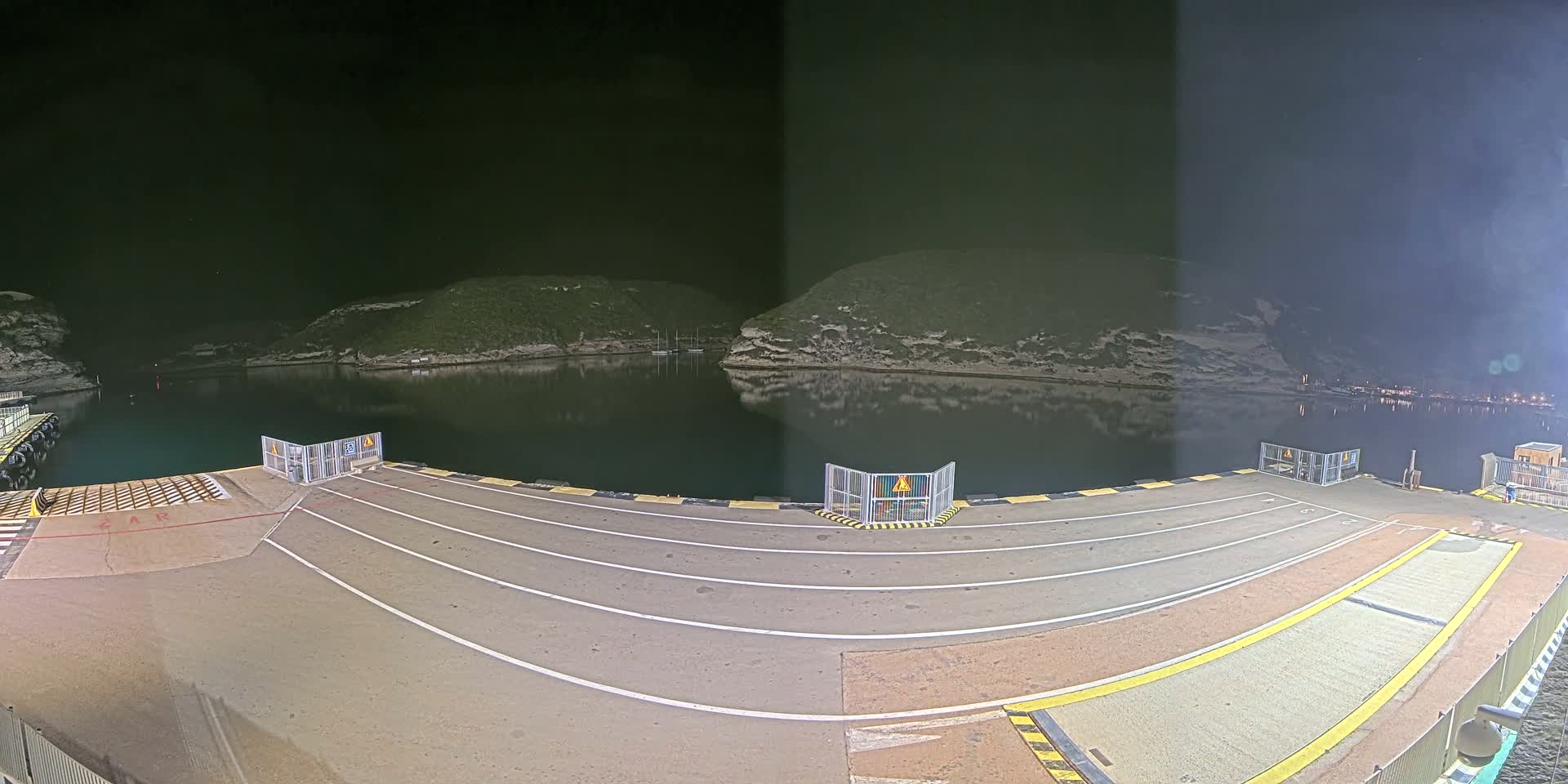 An empty ferry dock at night, illuminated, overlooks calm water reflecting the dark hillsides surrounding it.