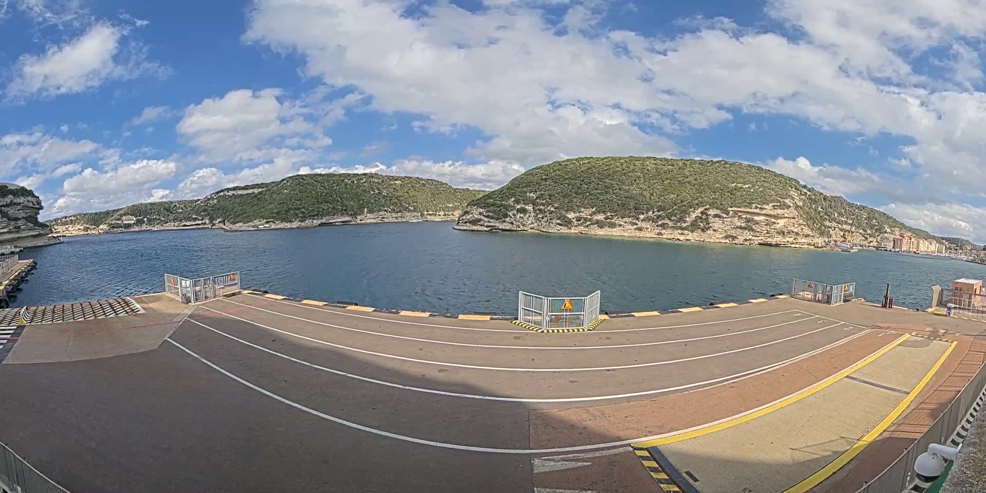 A ferry terminal's paved loading area overlooks a calm body of water surrounded by rocky, green hills under a partly cloudy sky.