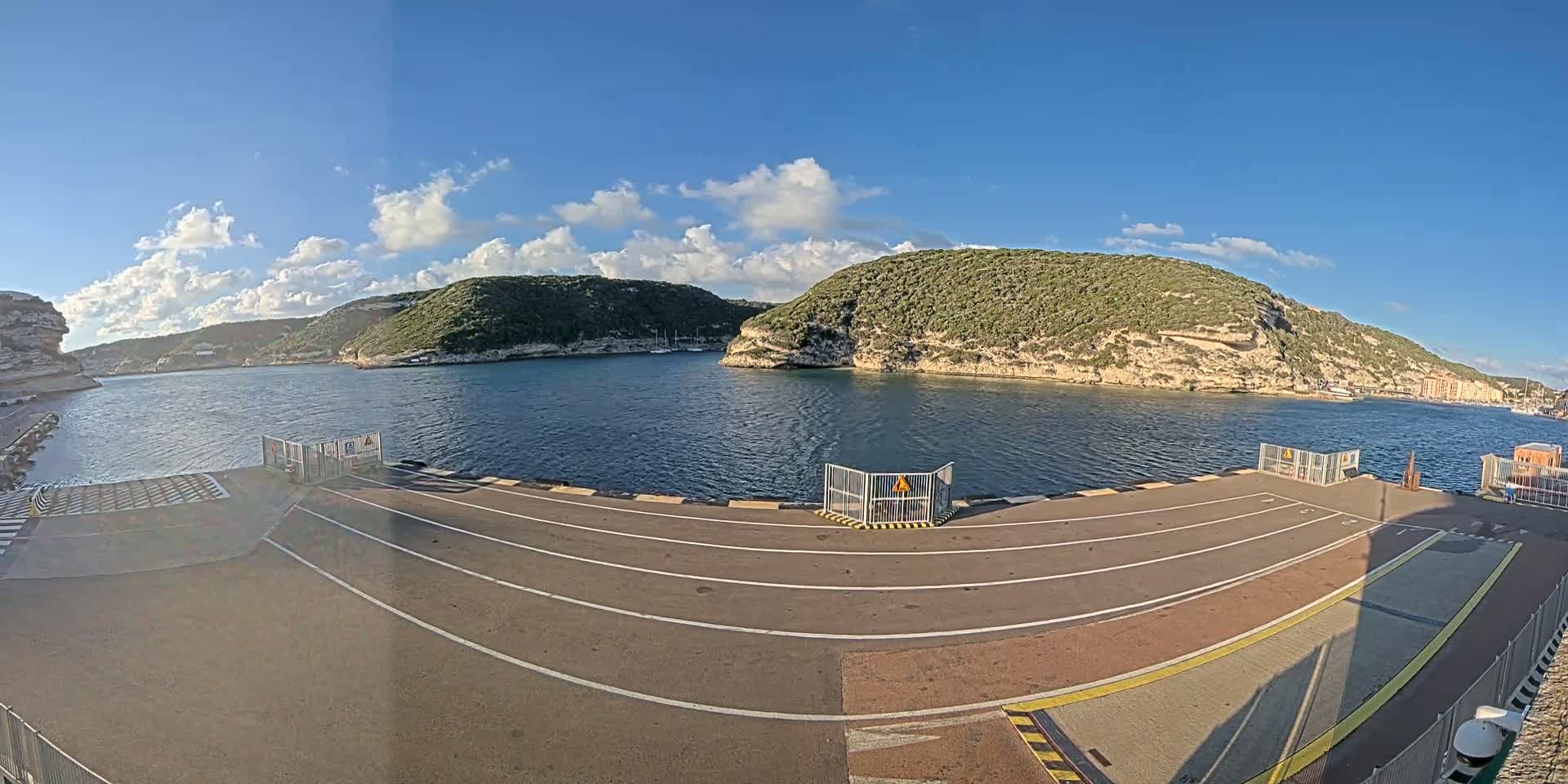 A paved ferry dock area overlooks a calm bay surrounded by green, rocky hills under a mostly sunny sky with some scattered clouds.