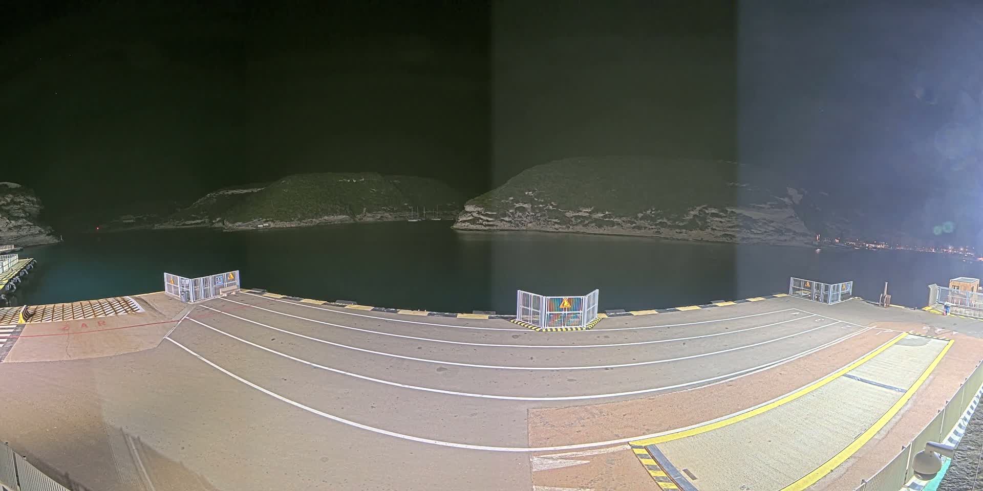 A nighttime view of an empty ferry dock overlooking a calm, dark body of water nestled between two hills.