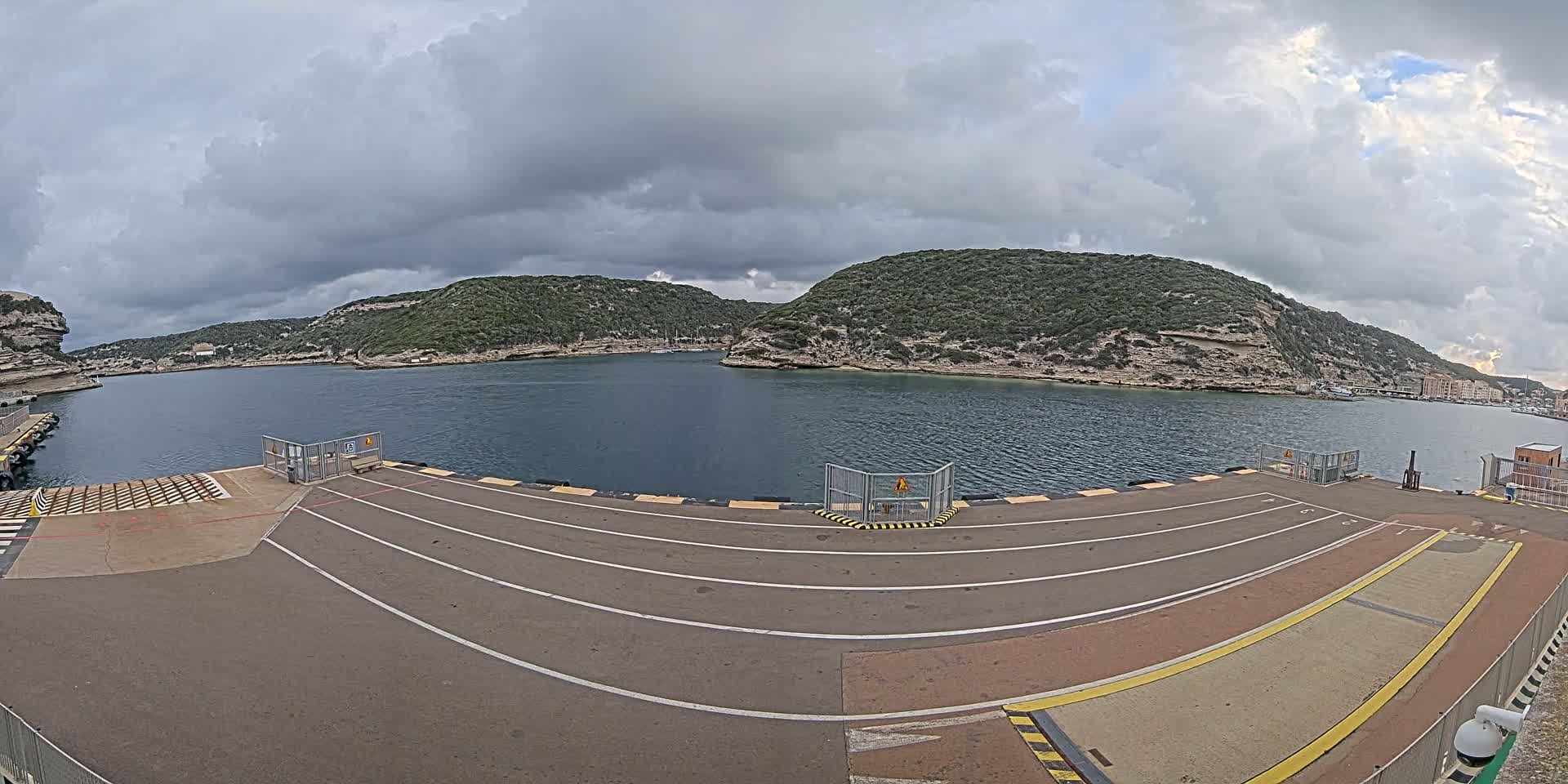 A ferry terminal with marked lanes overlooks a calm bay surrounded by green, rocky hills under a cloudy sky.