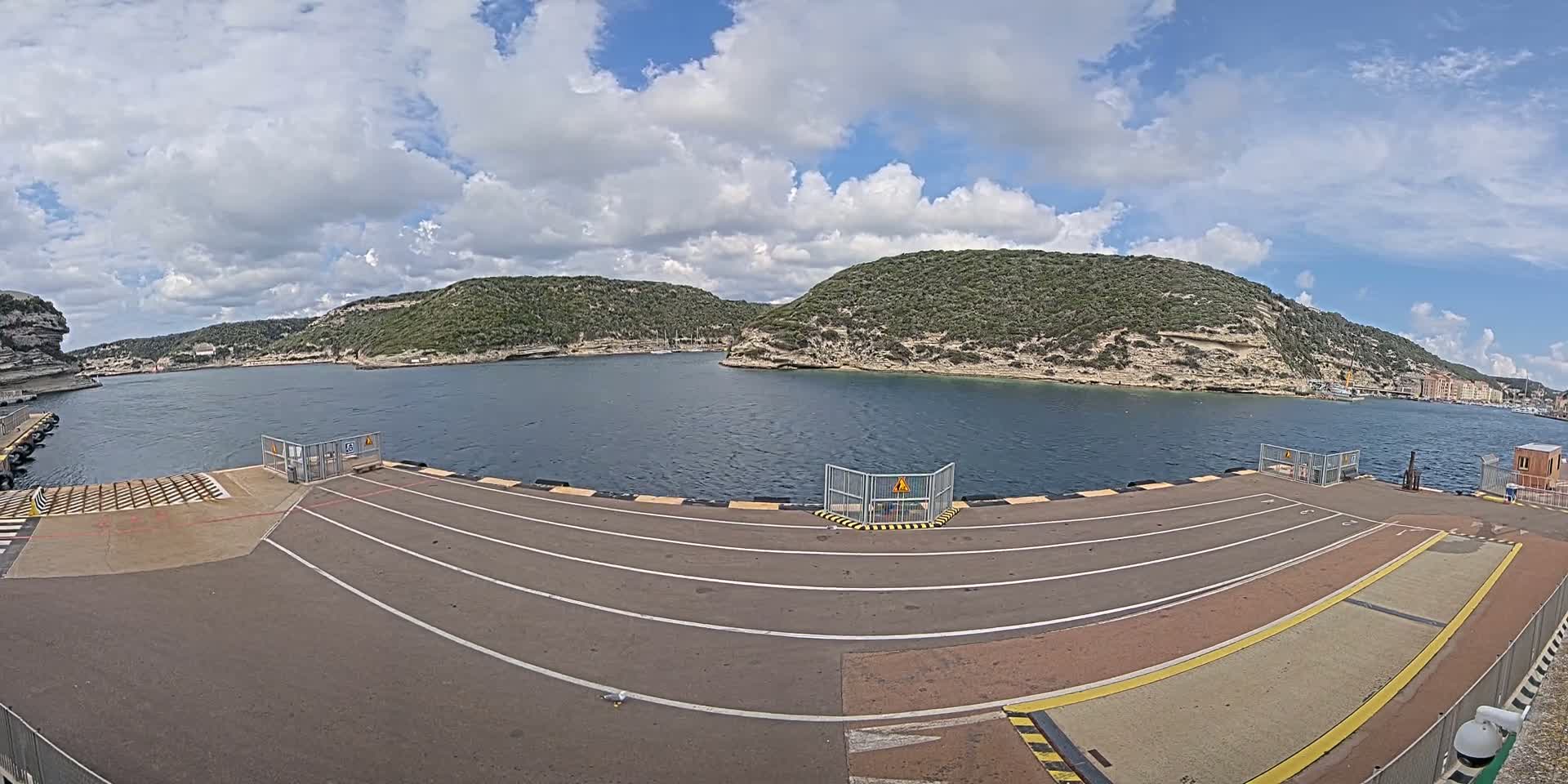 A ferry terminal with marked lanes overlooks a calm body of water surrounded by hilly, green terrain under a partly cloudy sky.