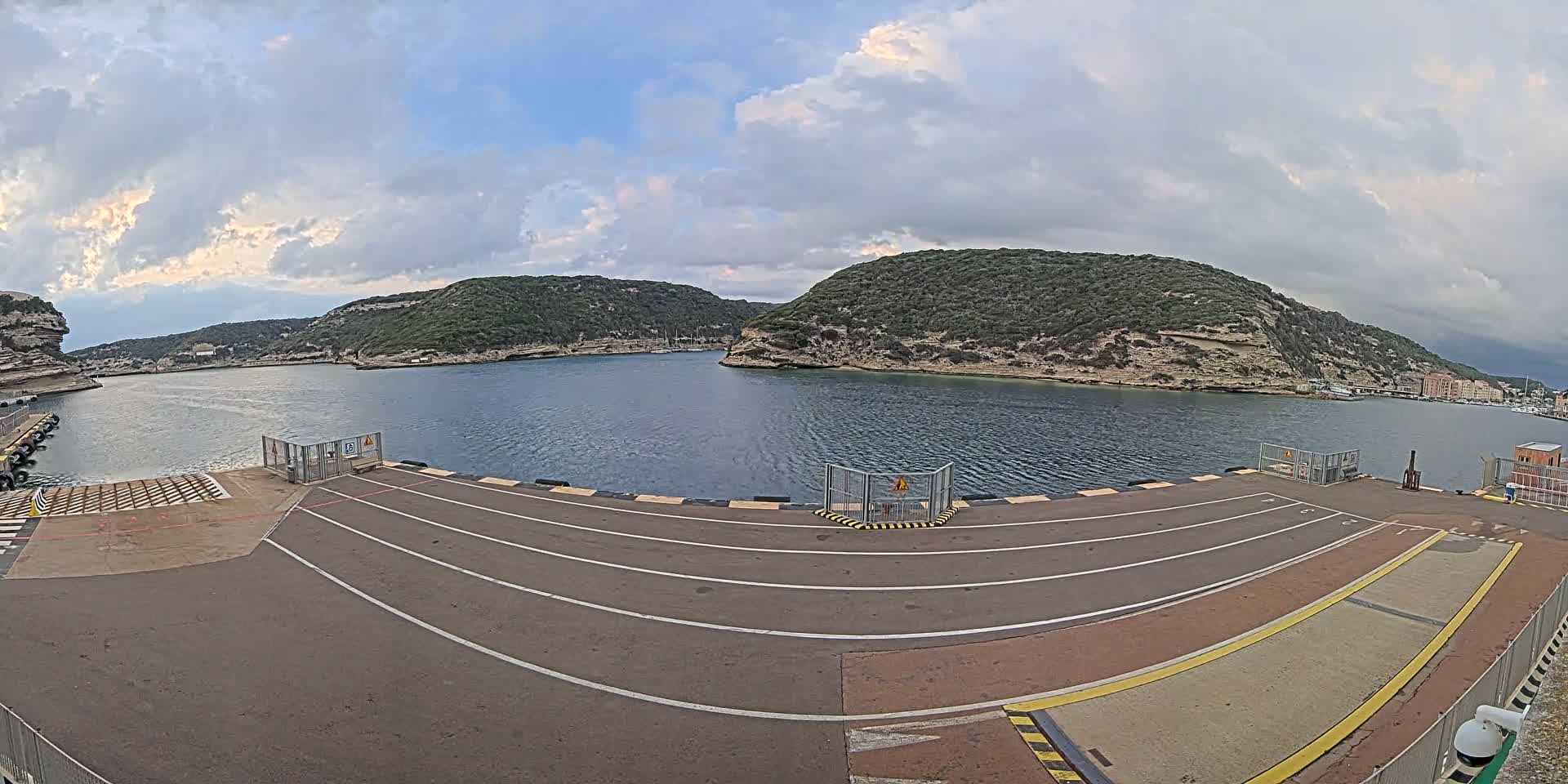 A ferry terminal with marked lanes overlooks a calm bay surrounded by green, rocky hills under a partly cloudy sky.