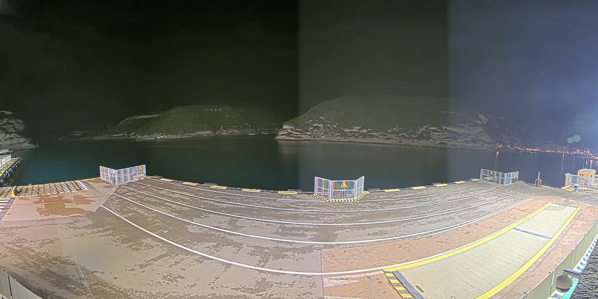 An empty ferry dock at night, with calm dark water and hills in the background under a dark sky.