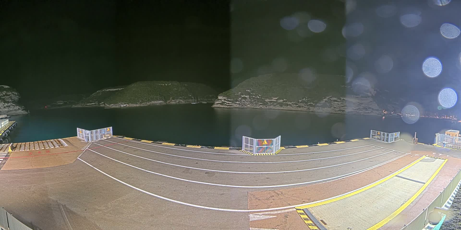 A nighttime view of an empty ferry dock with rain on the camera lens, overlooking a calm body of water surrounded by dark hills.