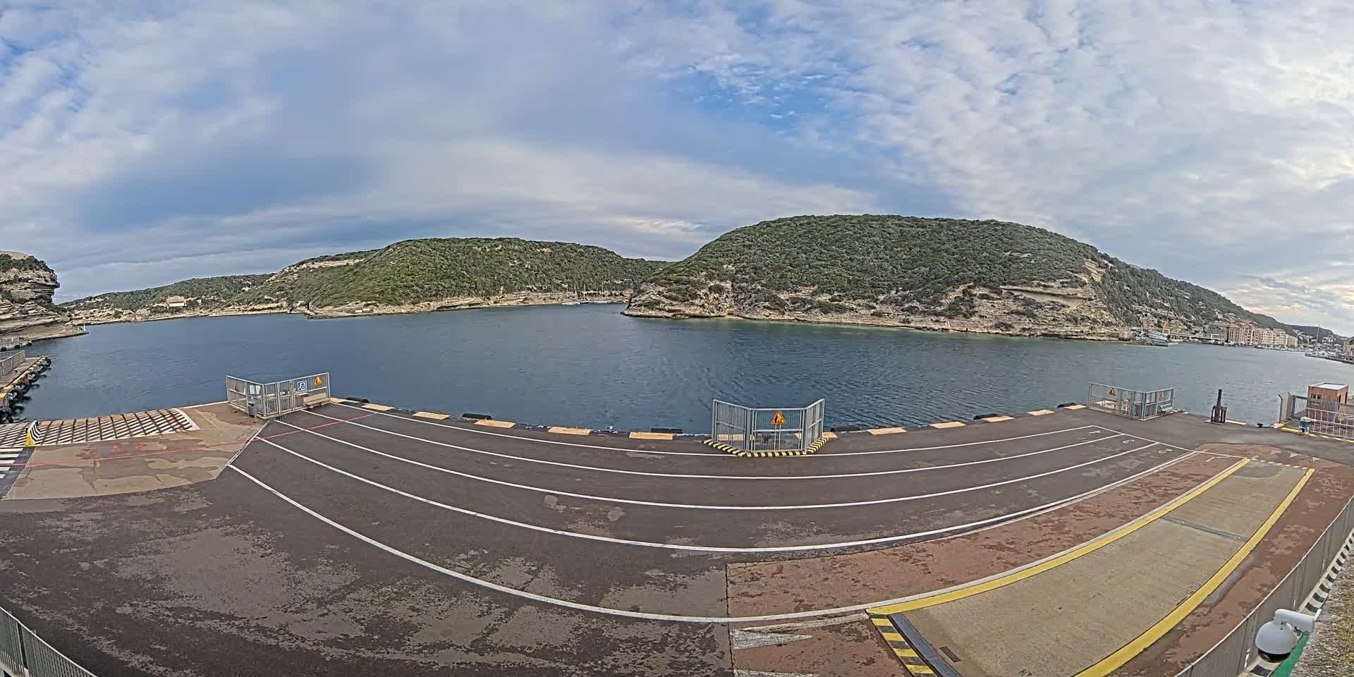 A ferry terminal's paved loading area overlooks a calm bay nestled between verdant, cliff-faced hills under a partly cloudy sky.