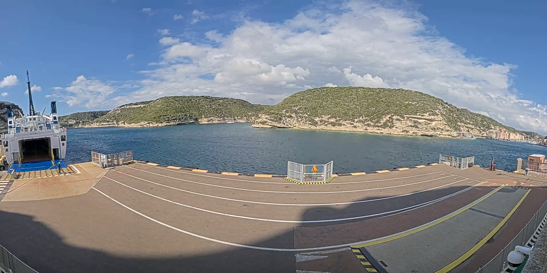 A ferry is docked at a port on a partly cloudy day, with calm water and green hills surrounding the bay.