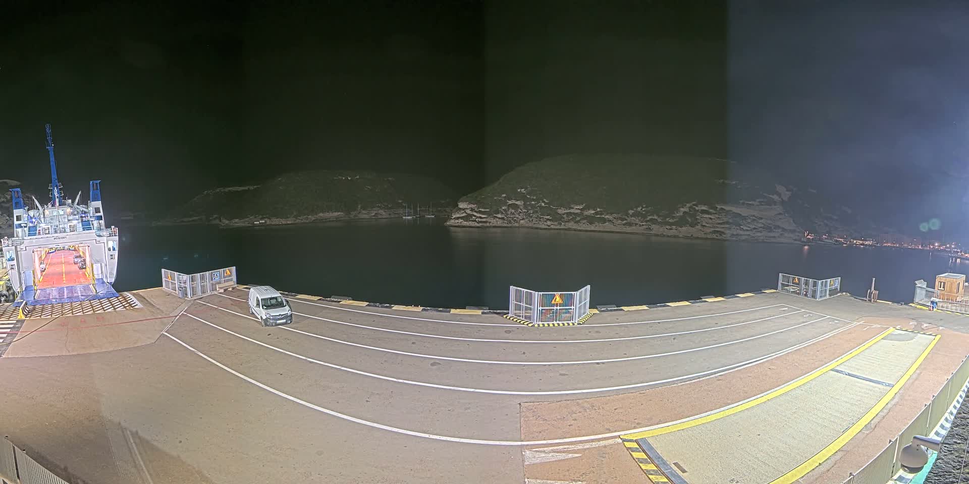 A ferry is docked at night at a port, with a van parked on the dock and calm water surrounding a small landmass.