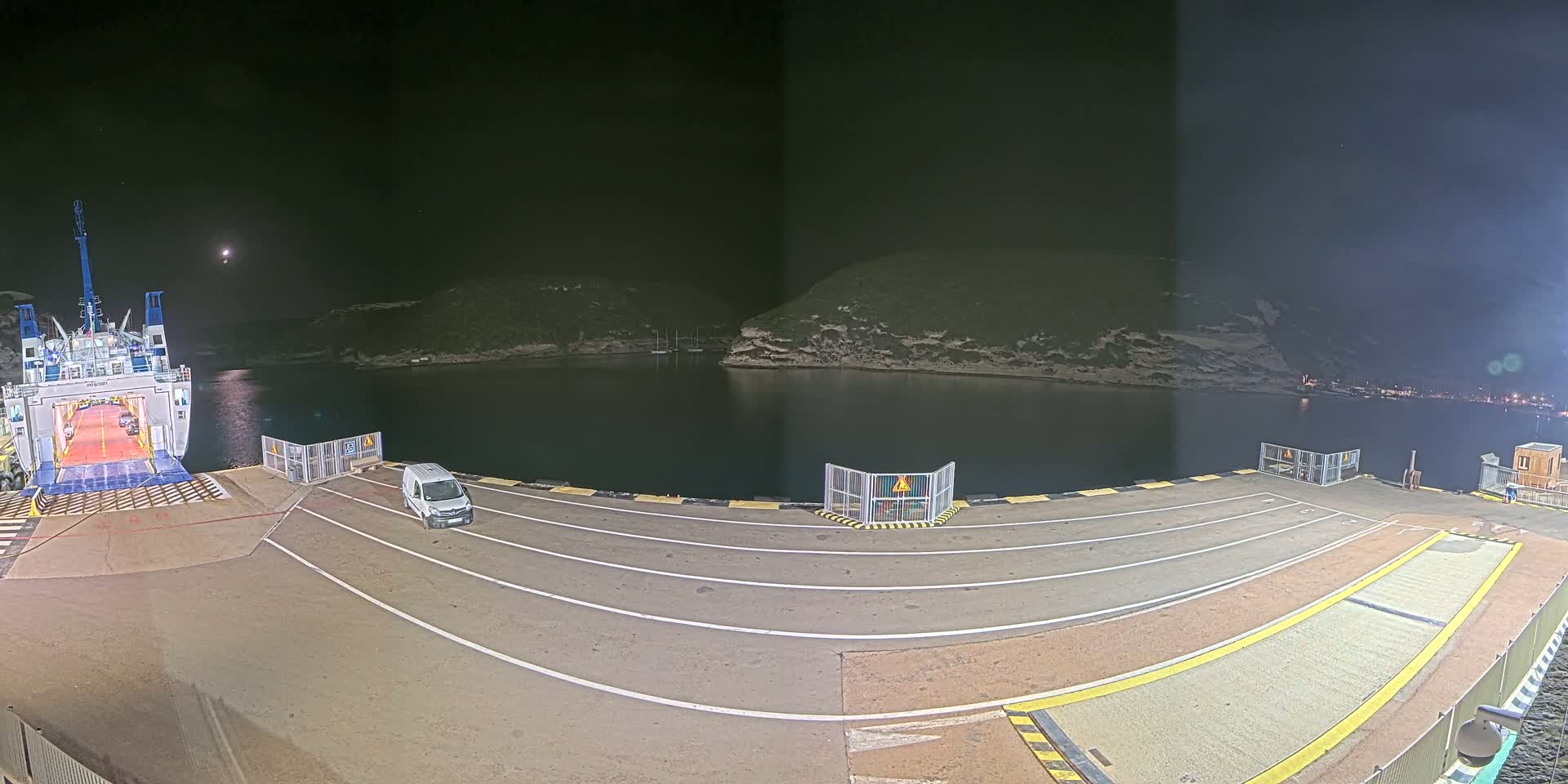 A ferry is docked at night in a calm harbor next to a van, under a partly cloudy night sky with a visible moon.