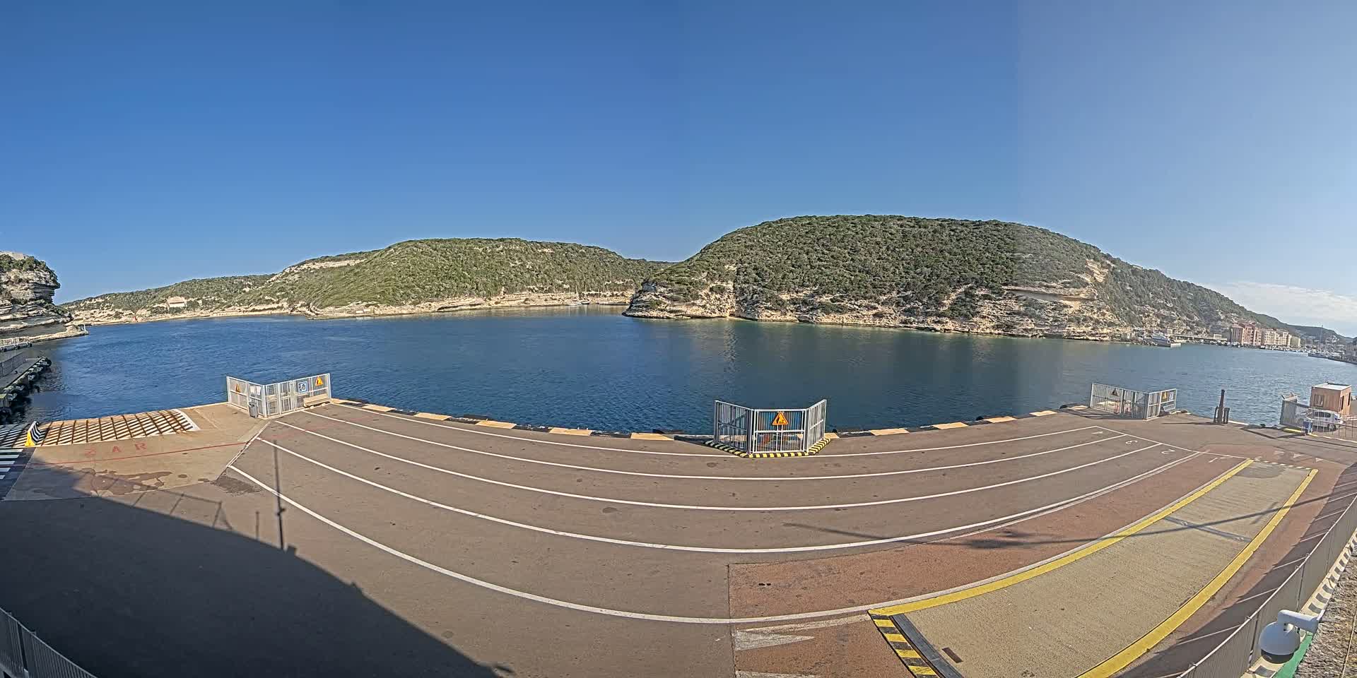 A ferry terminal's empty loading area overlooks a calm bay surrounded by green, rocky hills under a clear blue sky.