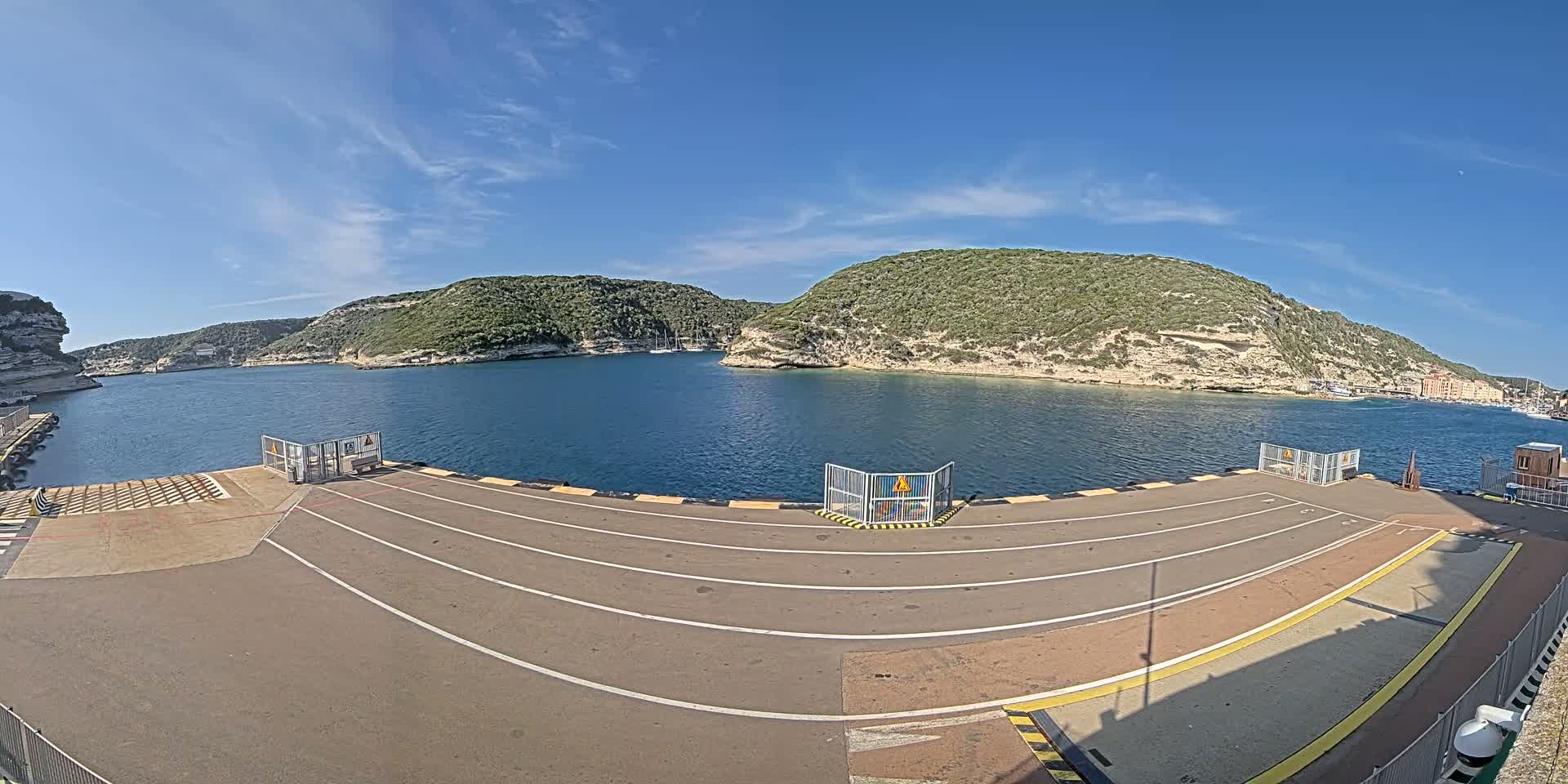 A ferry dock with marked lanes overlooks a calm bay bordered by green, rocky hills under a clear blue sky.