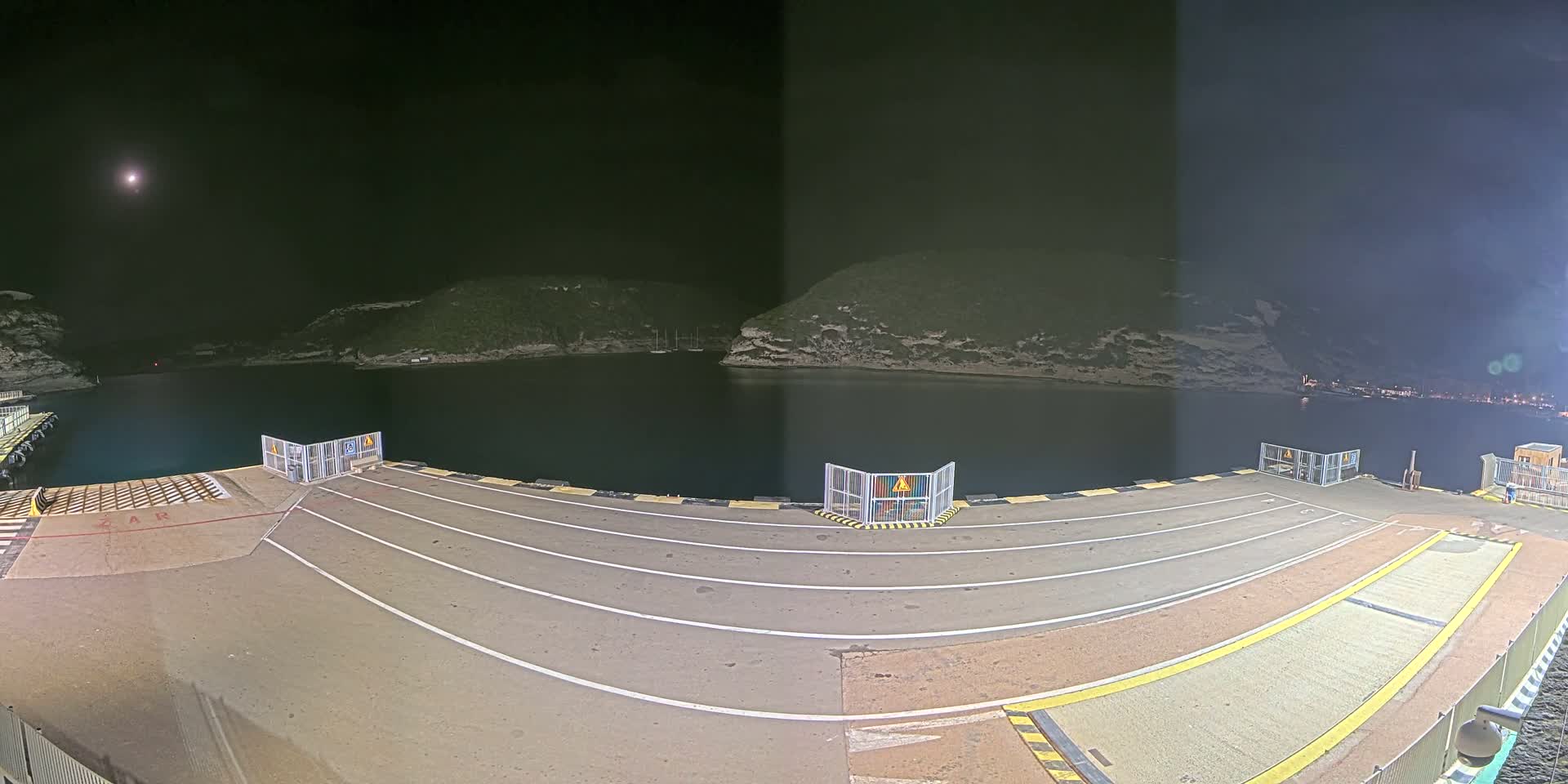 An empty ferry dock at night under a clear sky with a bright moon is illuminated, overlooking calm dark water and hills in the distance.