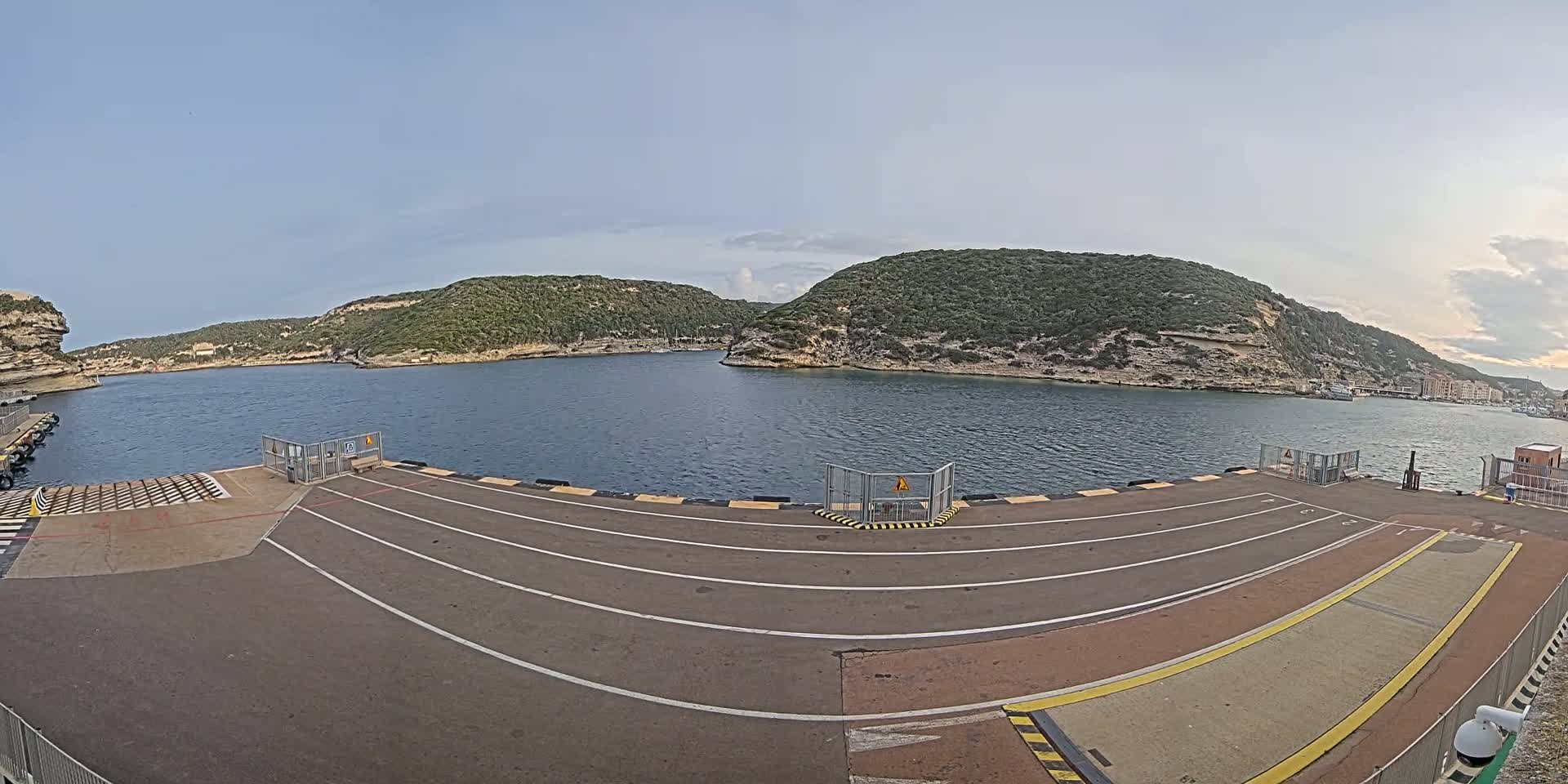 An empty ferry dock with marked lanes overlooks a calm bay surrounded by green, rocky hills under a mostly clear sky.