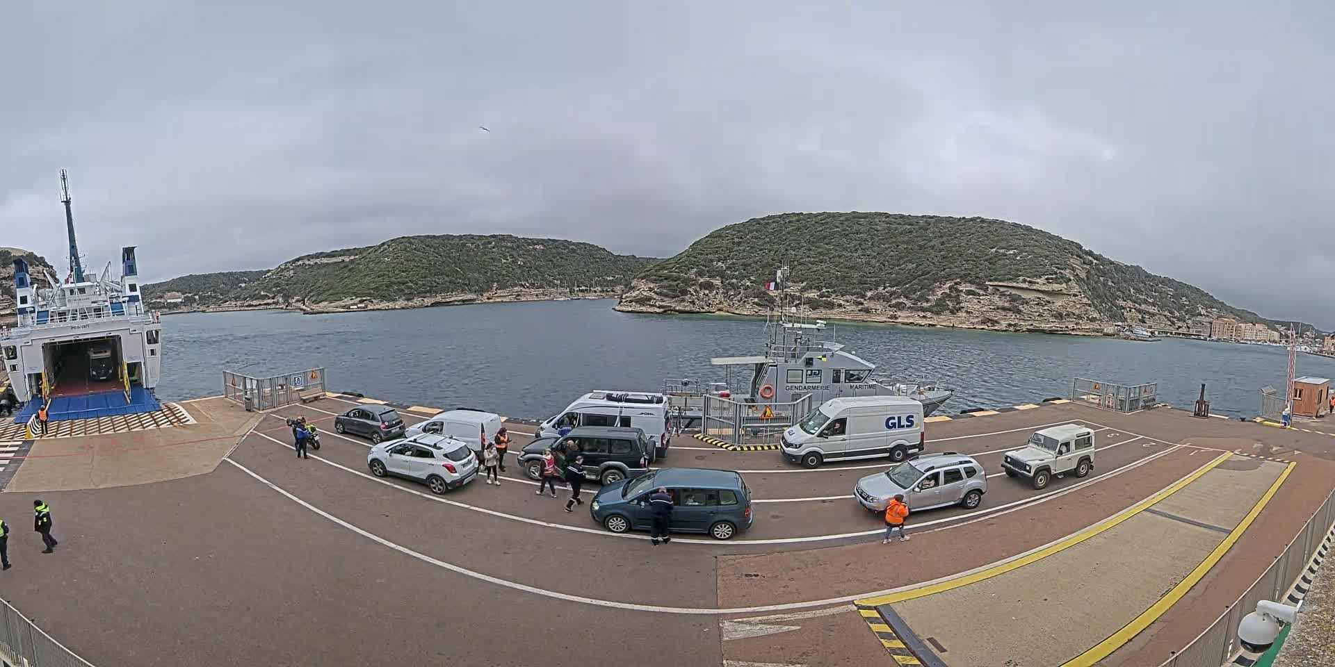 A ferry is docked at a port, with several vehicles and people on a paved area, under an overcast sky.