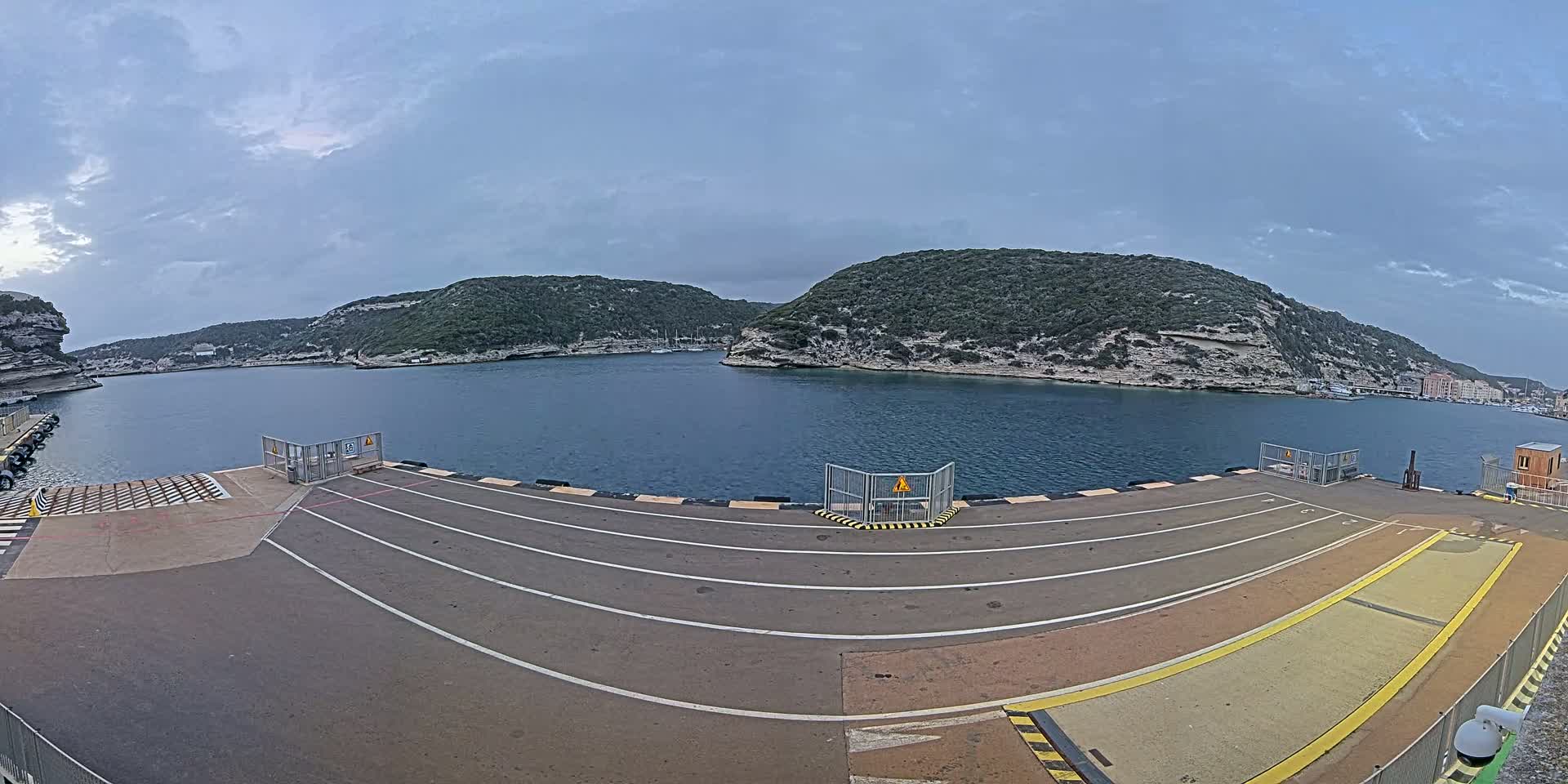 An empty ferry dock with marked lanes overlooks a calm body of water surrounded by green hills under a cloudy sky.