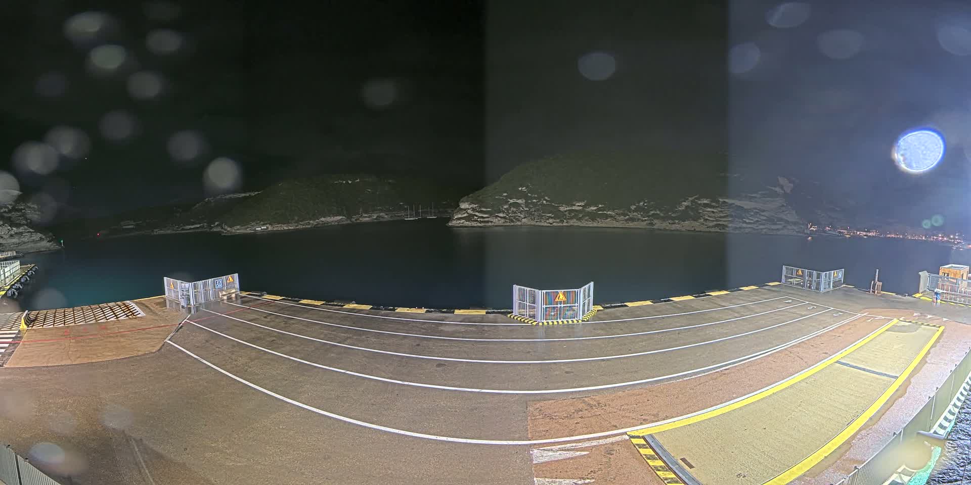 A nighttime view of a ferry terminal with rain, showing an empty dock area and a calm body of water surrounded by dark hills under a mostly clear sky.