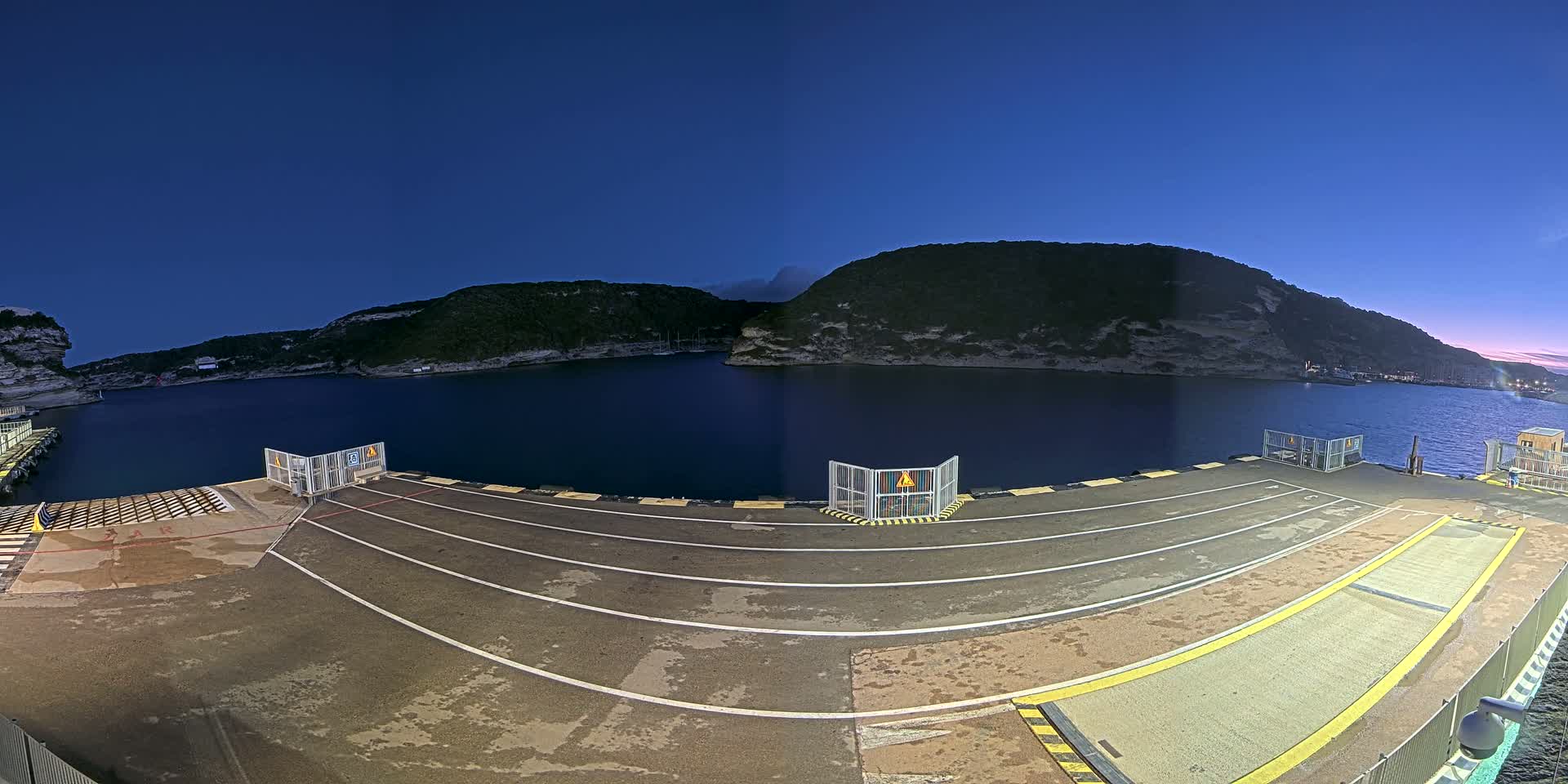 An empty ferry dock at night, under a clear, dark blue sky, overlooks calm water surrounded by hills.